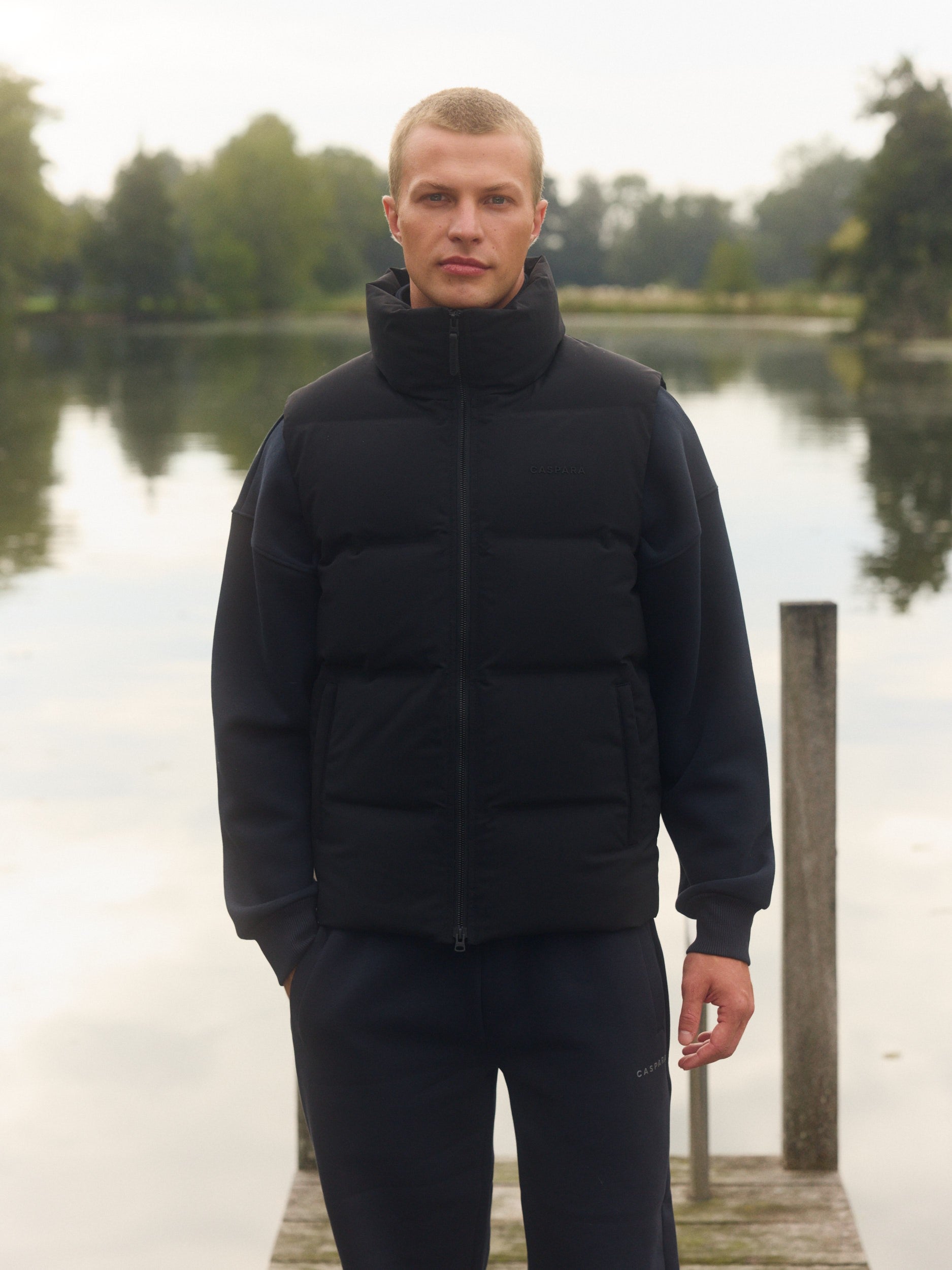Man wearing a black puffer vest standing on a wooden dock by a lake.