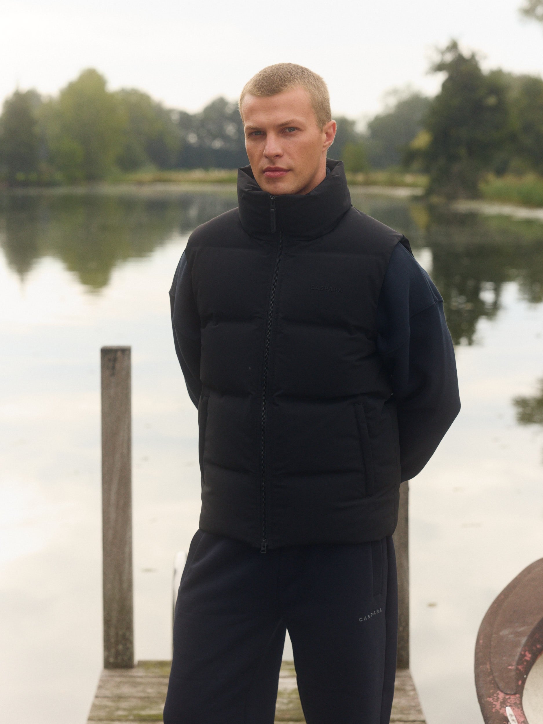 Man wearing a matte black puffer vest standing on a wooden dock by a lake.