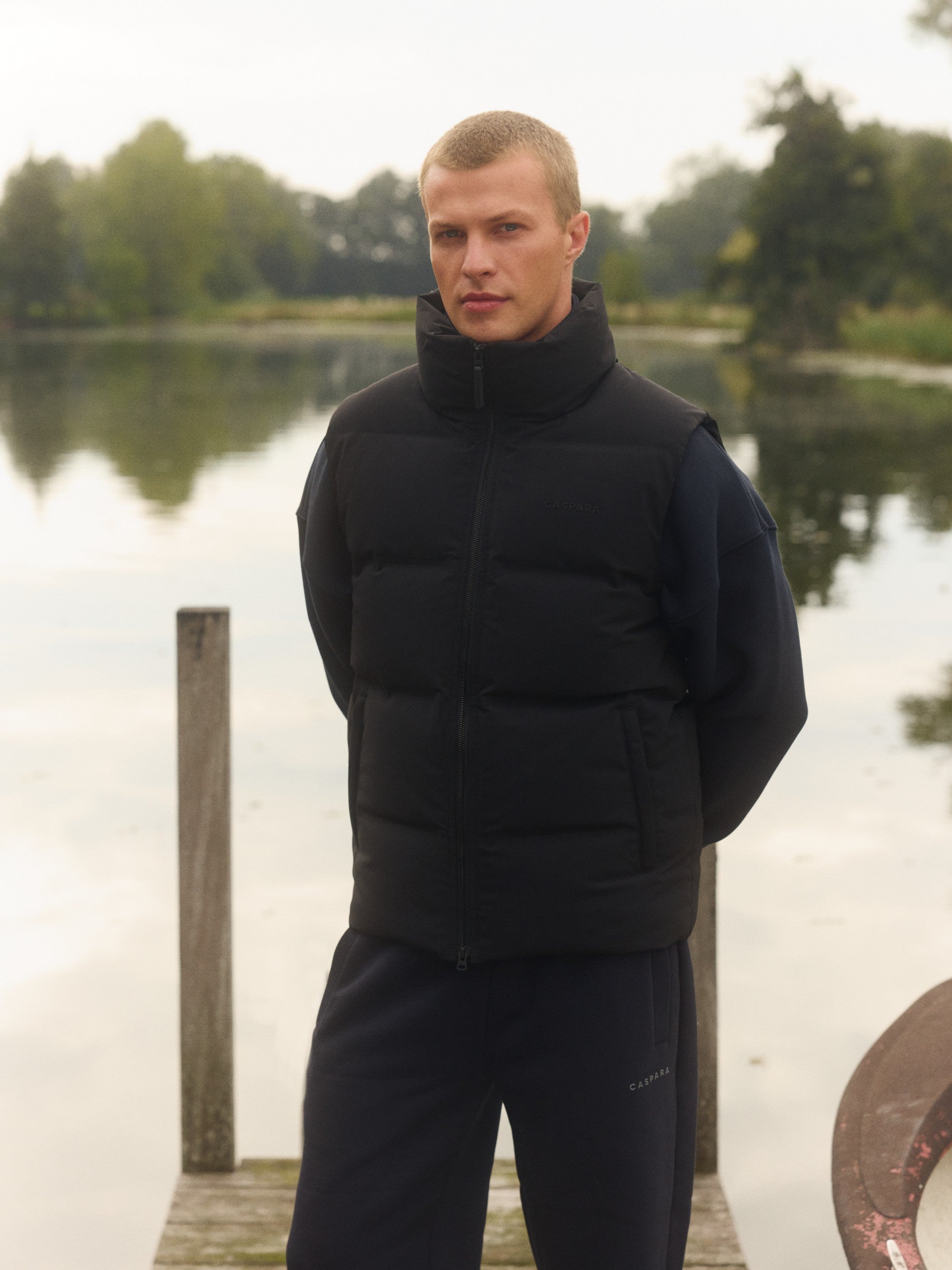 Man wearing a black CASPARA Boreal puffer vest on a wooden dock by a lake.