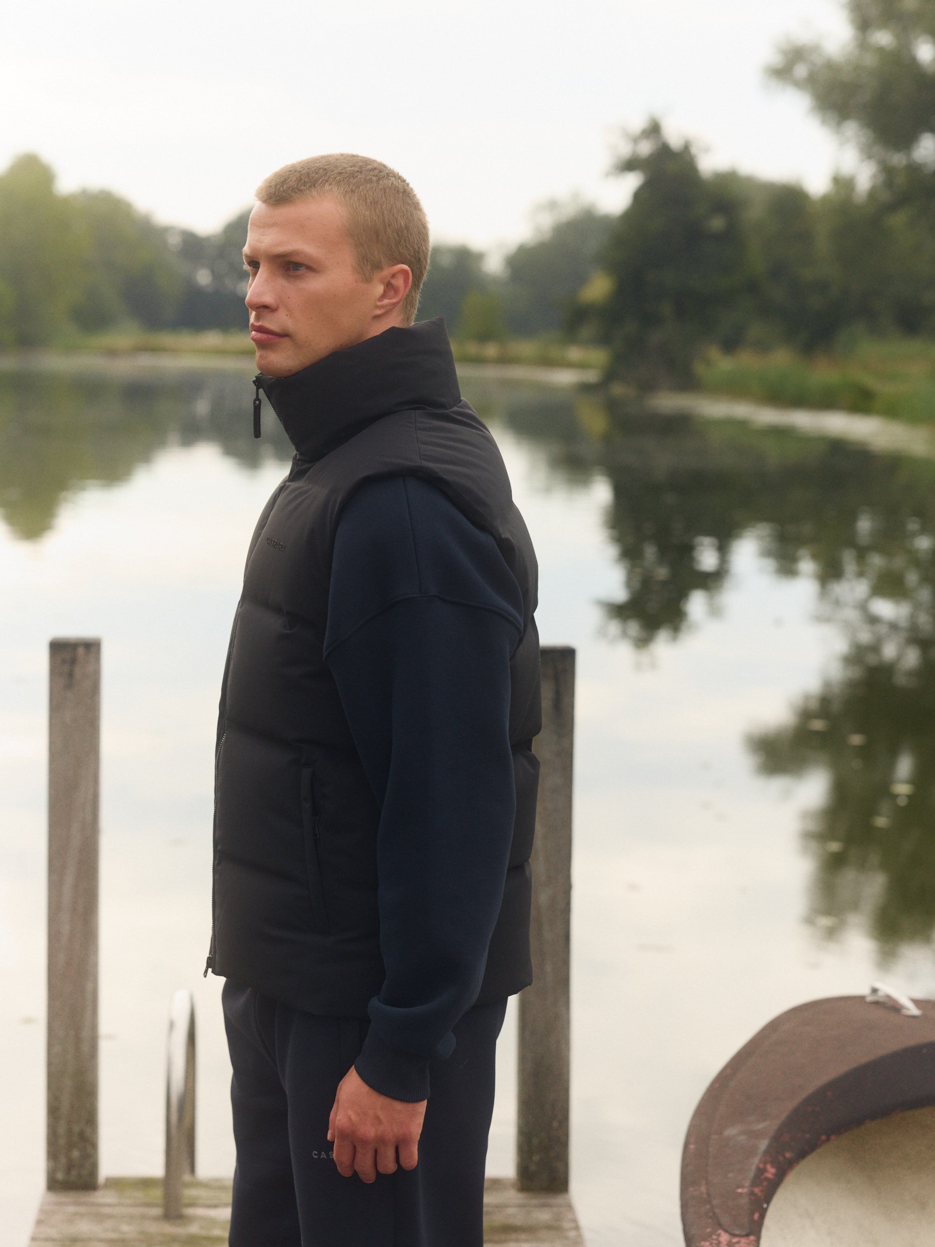 Man wearing a black matte puffer vest standing outdoors on a wooden dock by a lake.