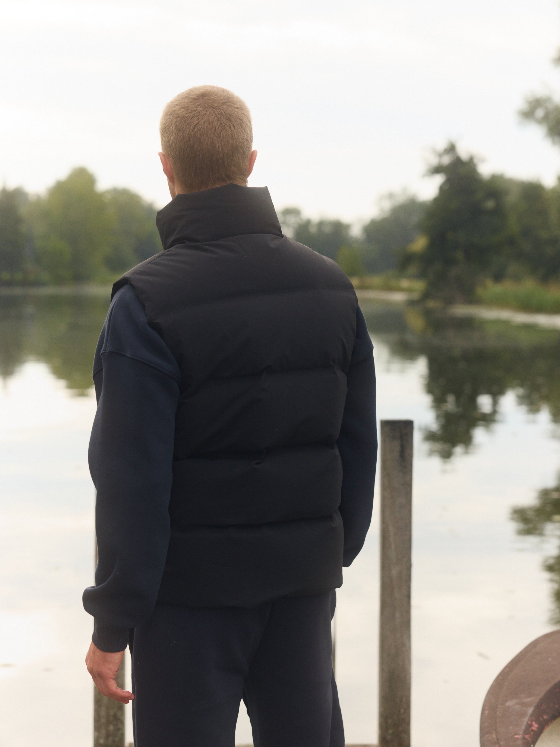 Man seen from behind wearing a black puffer vest standing on a wooden dock beside a lake.
