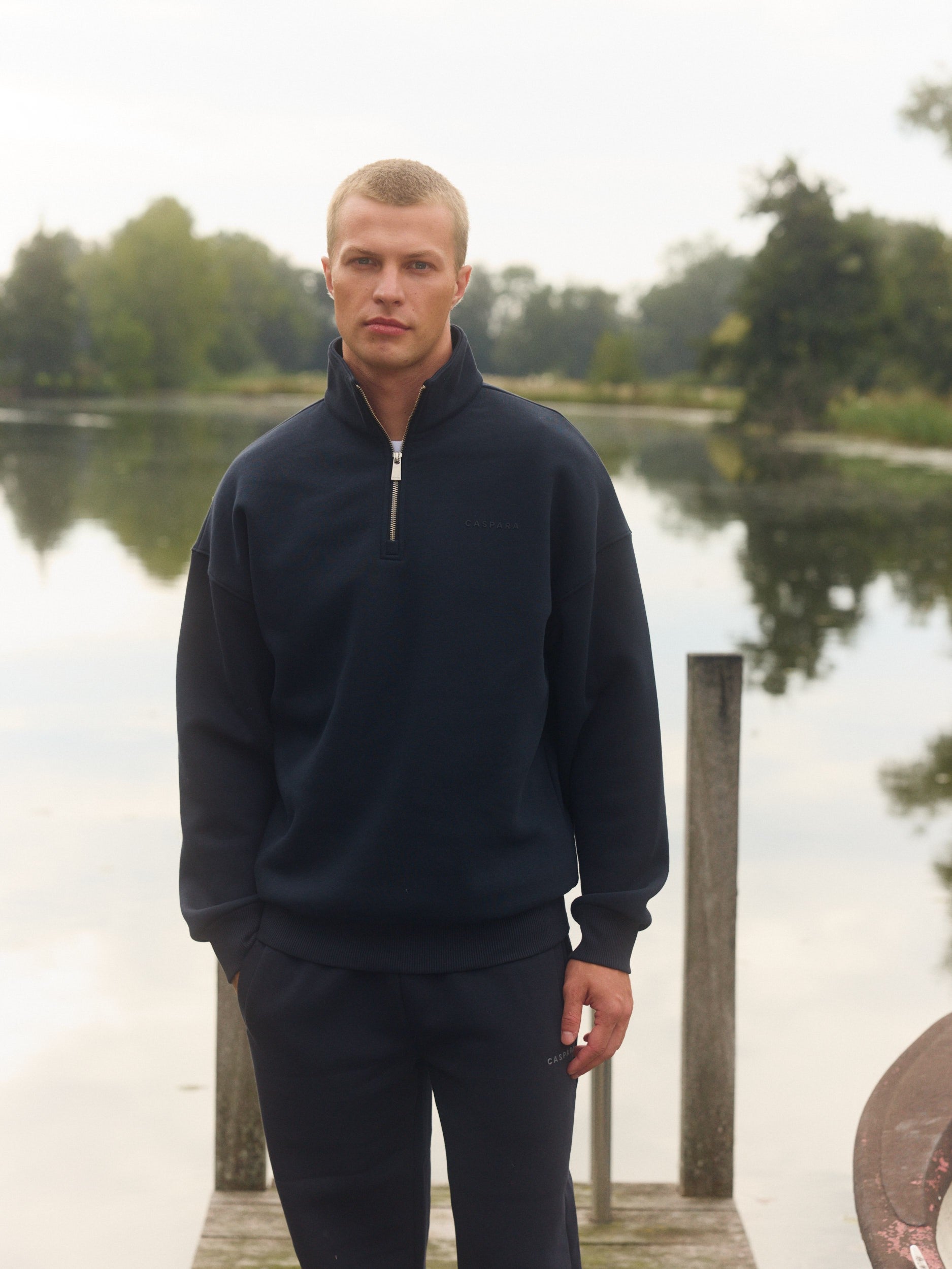 Man wearing a navy blue quarter-zip sweatshirt standing on a wooden dock by a lake.
