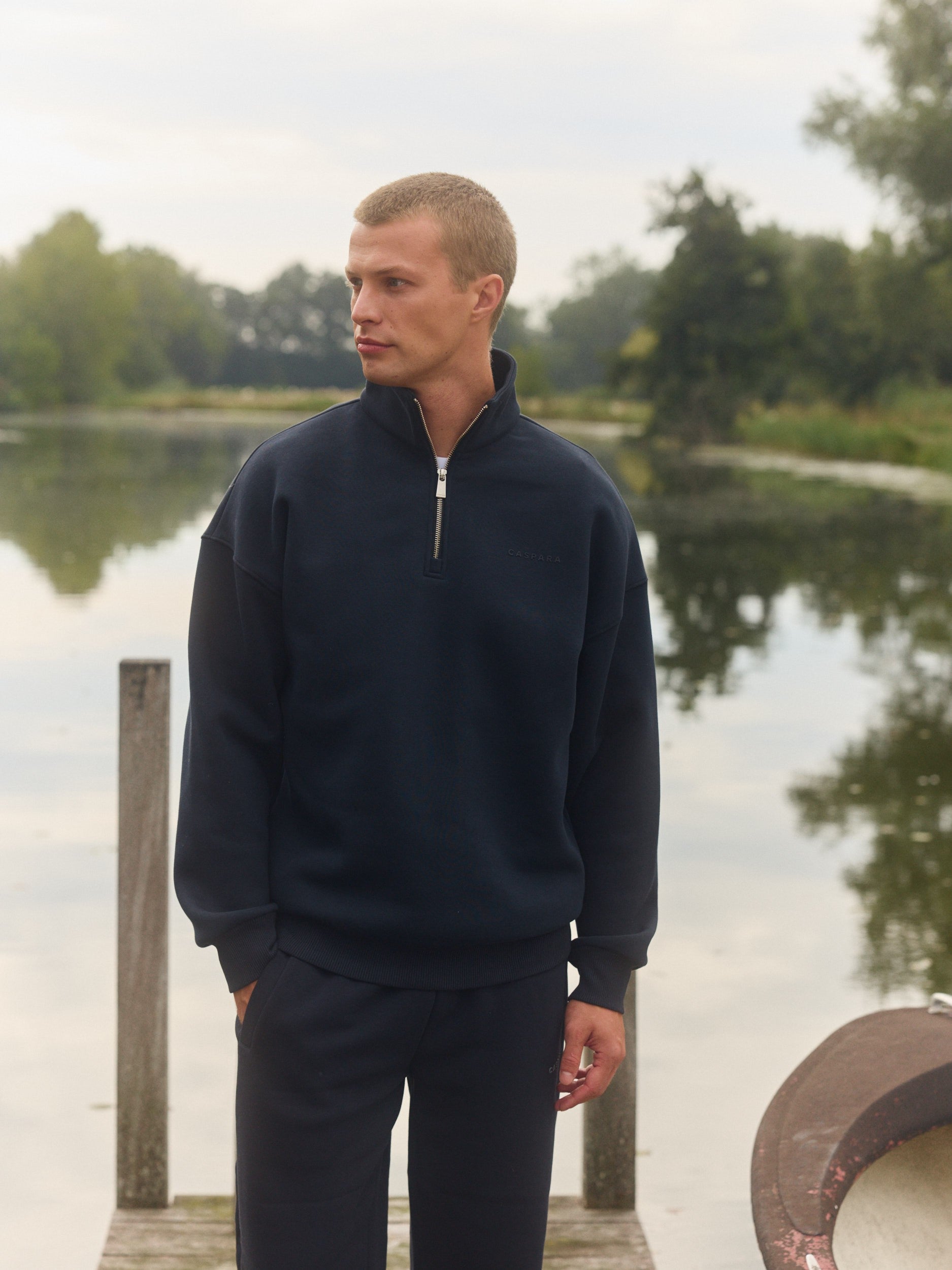Man wearing a navy quarter-zip sweatshirt standing on a wooden dock by a lake.
