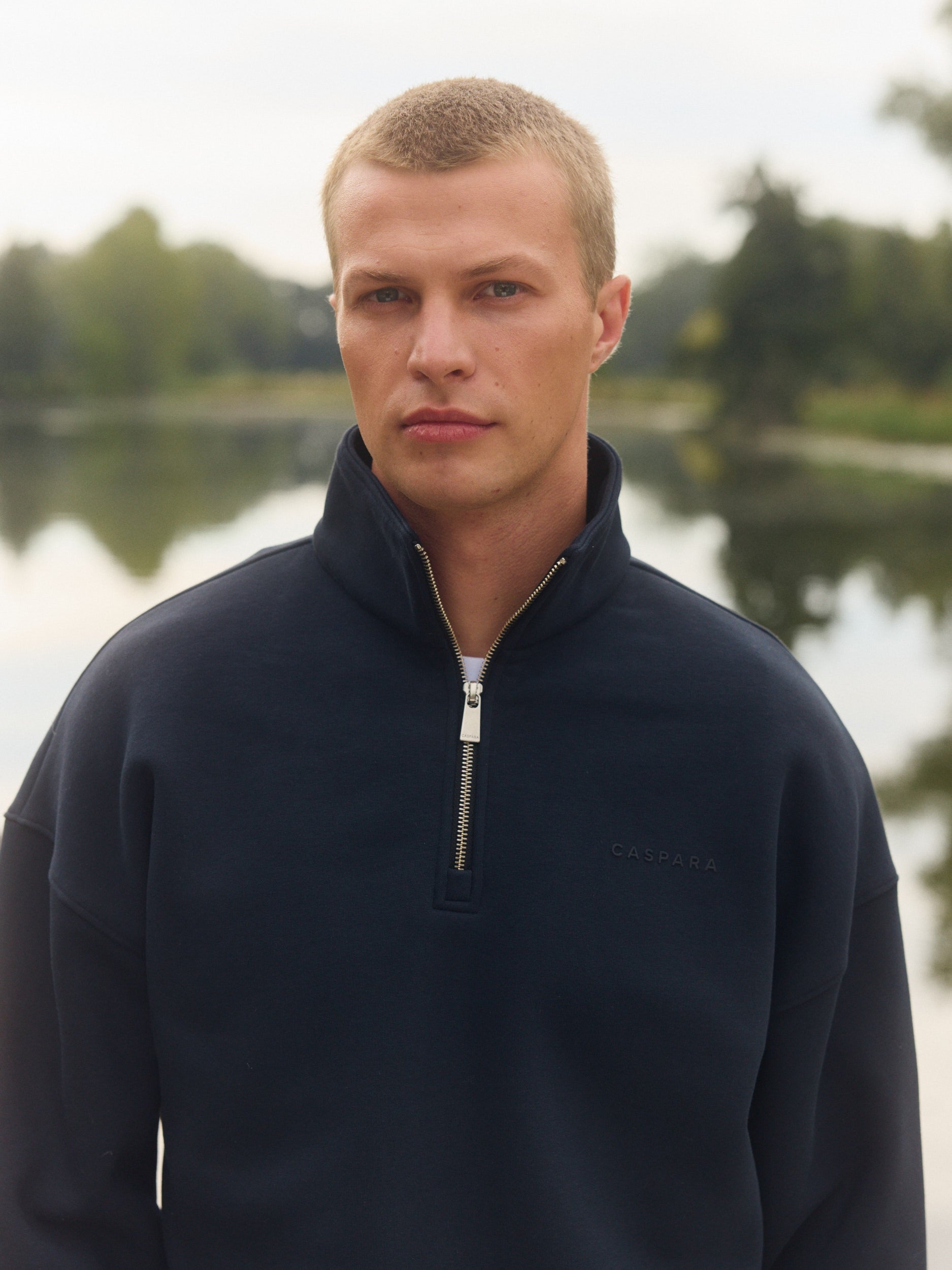 Man wearing a navy blue quarter-zip sweatshirt outdoors by a lake.