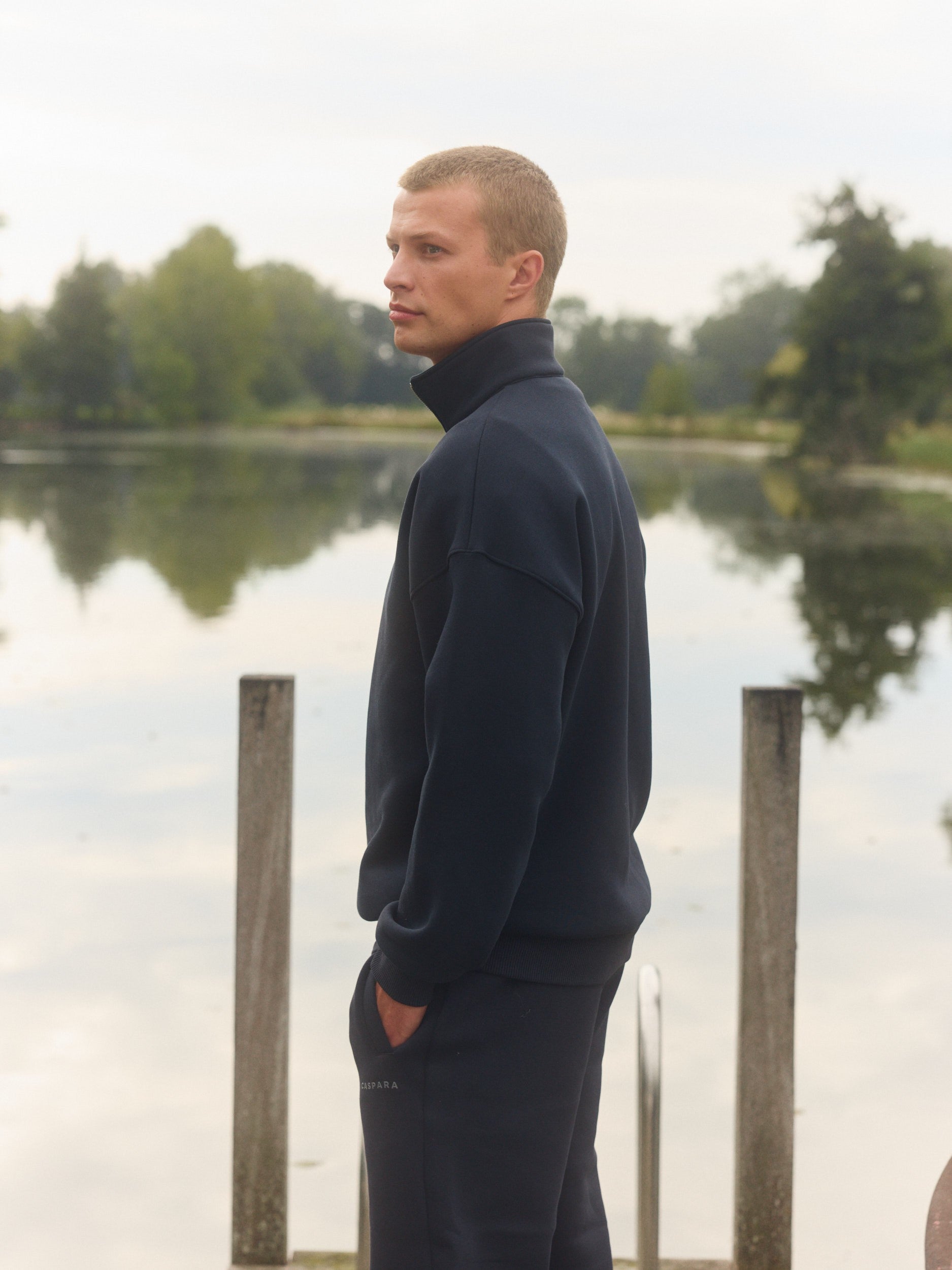 Man wearing a navy quarter-zip sweatshirt standing on a wooden dock by a lake.