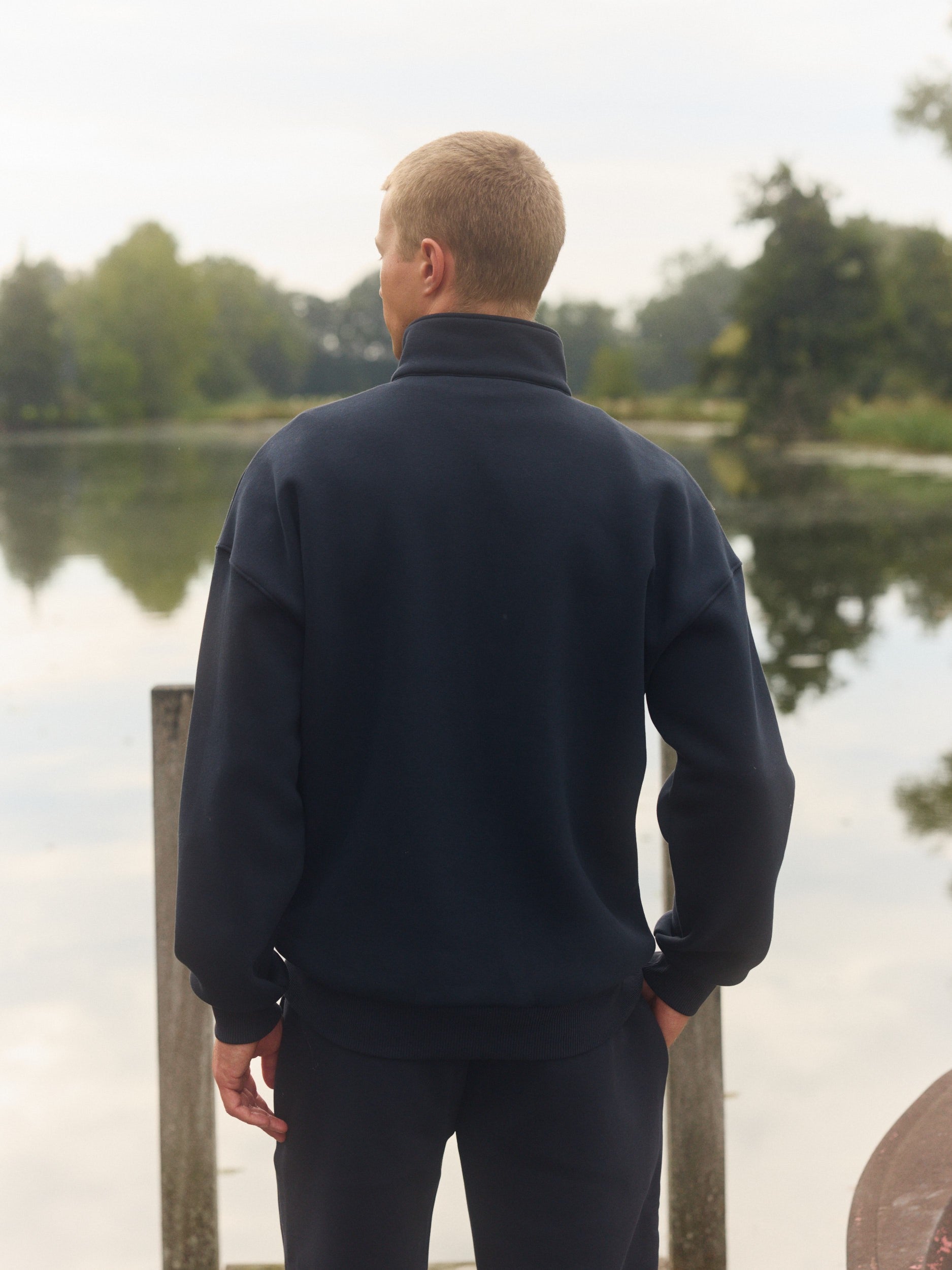 Back view of a man wearing a navy quarter-zip sweatshirt standing by a calm lake.