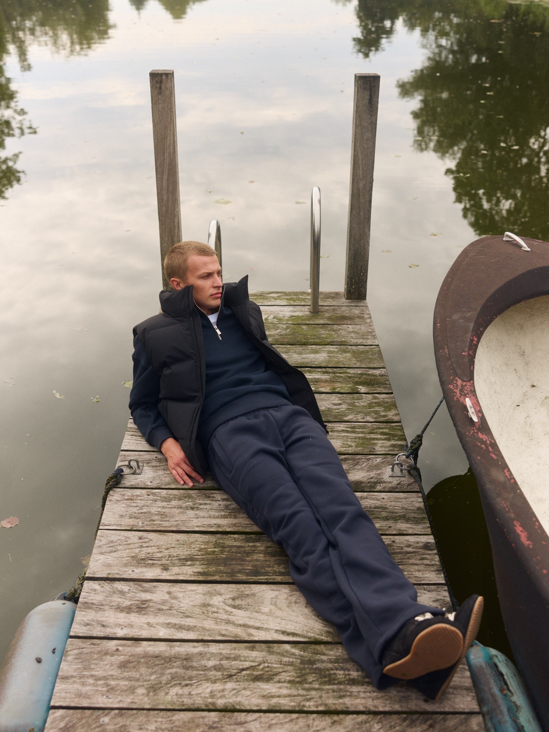 Man wearing a black puffer vest over a navy sweater lounging on a wooden dock by the water.