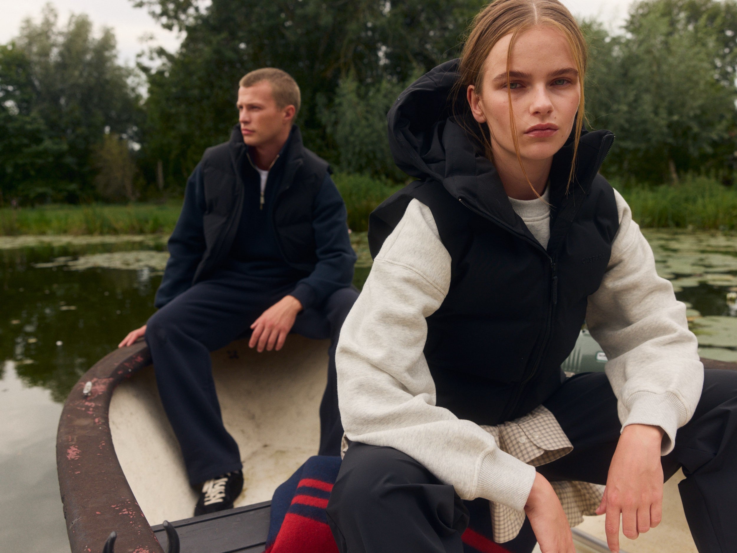 Woman and man wearing black hooded puffer vests sitting in a boat on a lake.