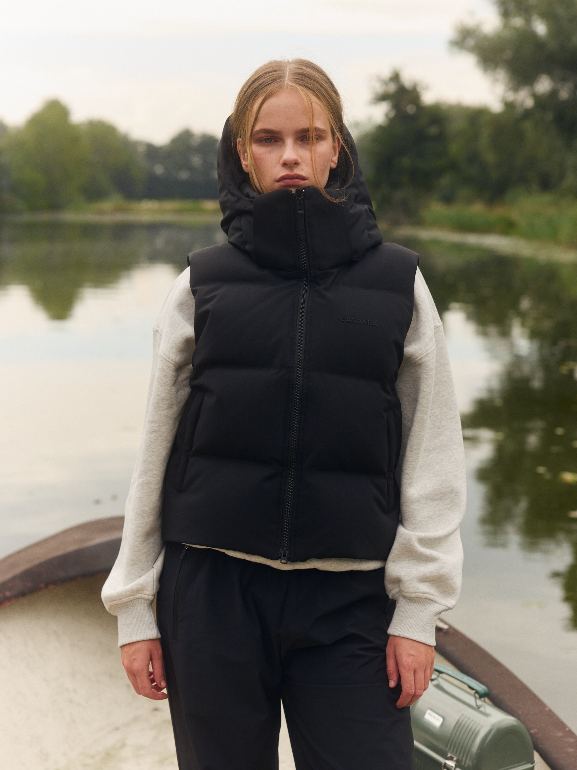 Woman wearing a black hooded puffer vest over a grey sweatshirt outdoors by a lake.