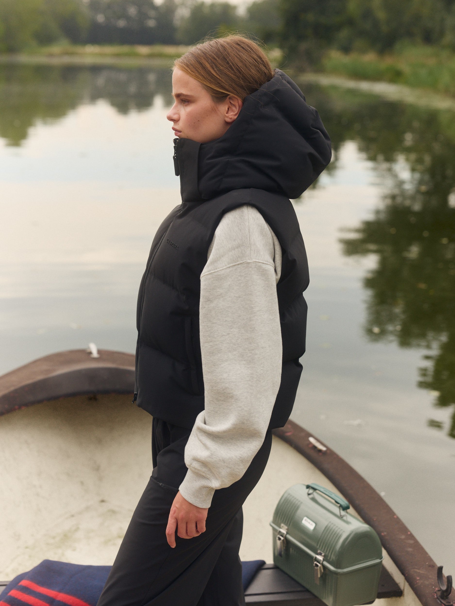 Woman wearing a black hooded puffer vest over a grey sweatshirt on a boat by a lake.