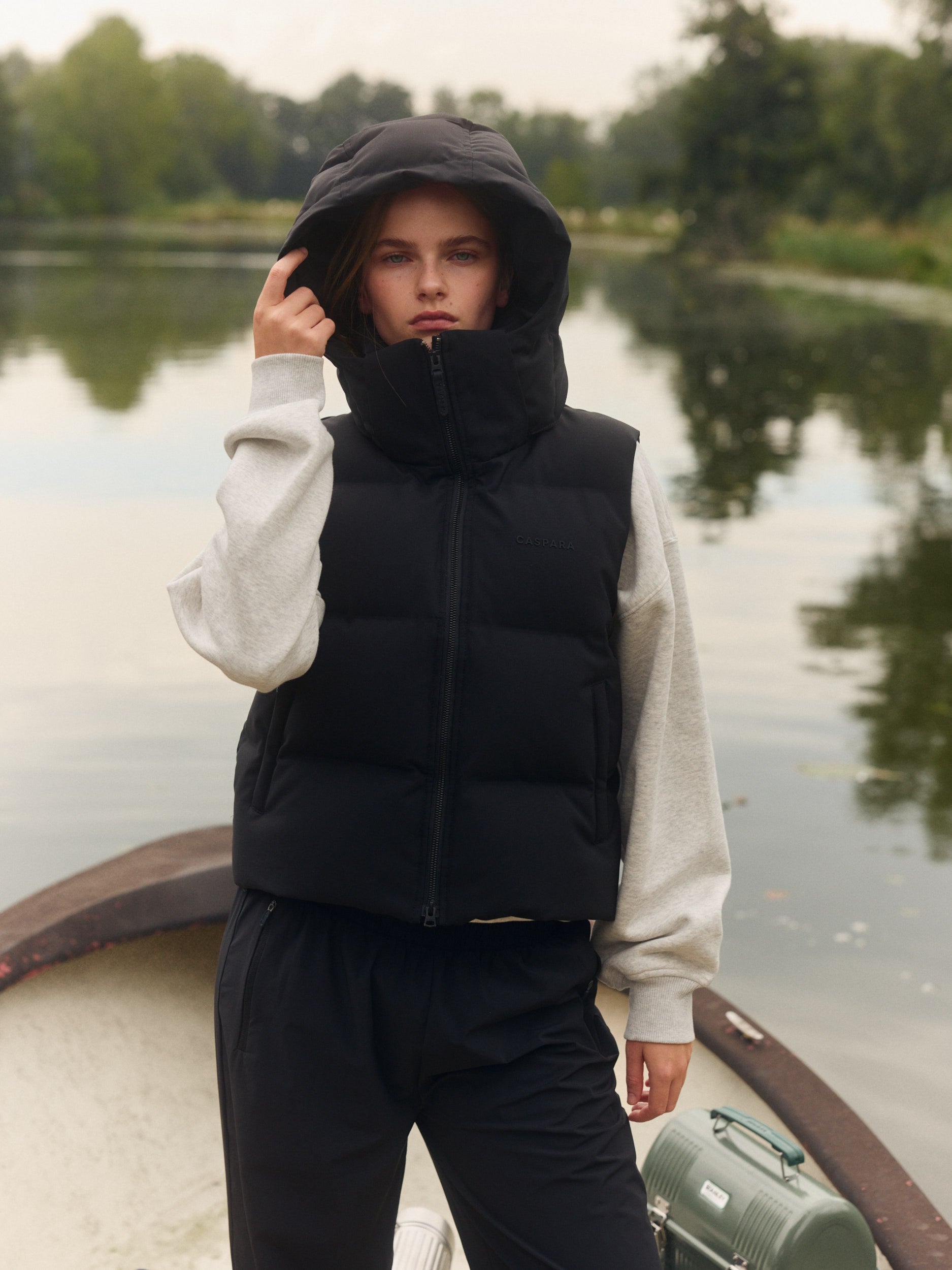 Woman wearing a black hooded puffer vest over a grey sweatshirt outdoors by a lake.