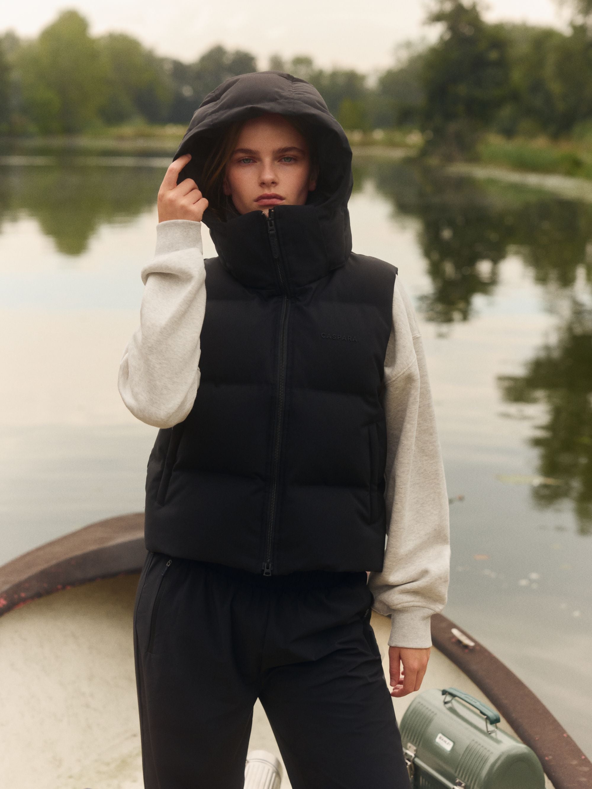 Woman wearing a black CASPARA hooded puffer vest outdoors by the water.