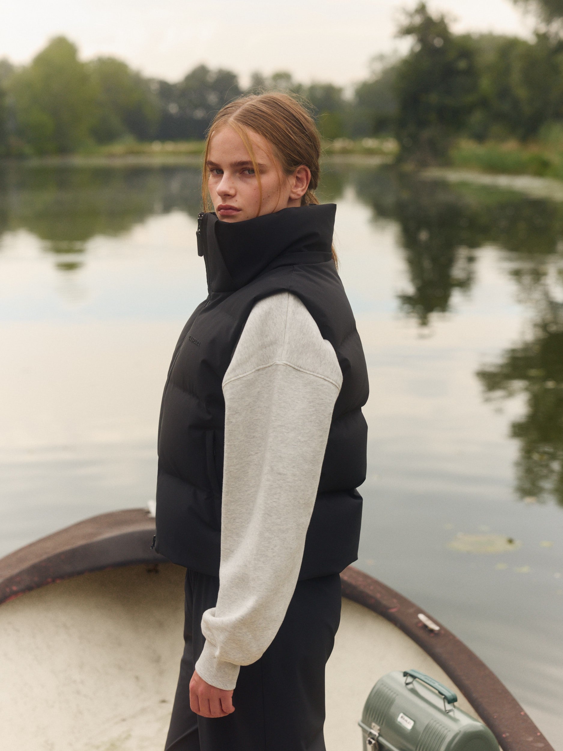 Woman wearing a black puffer vest over a grey sweatshirt standing in a boat on a calm lake.