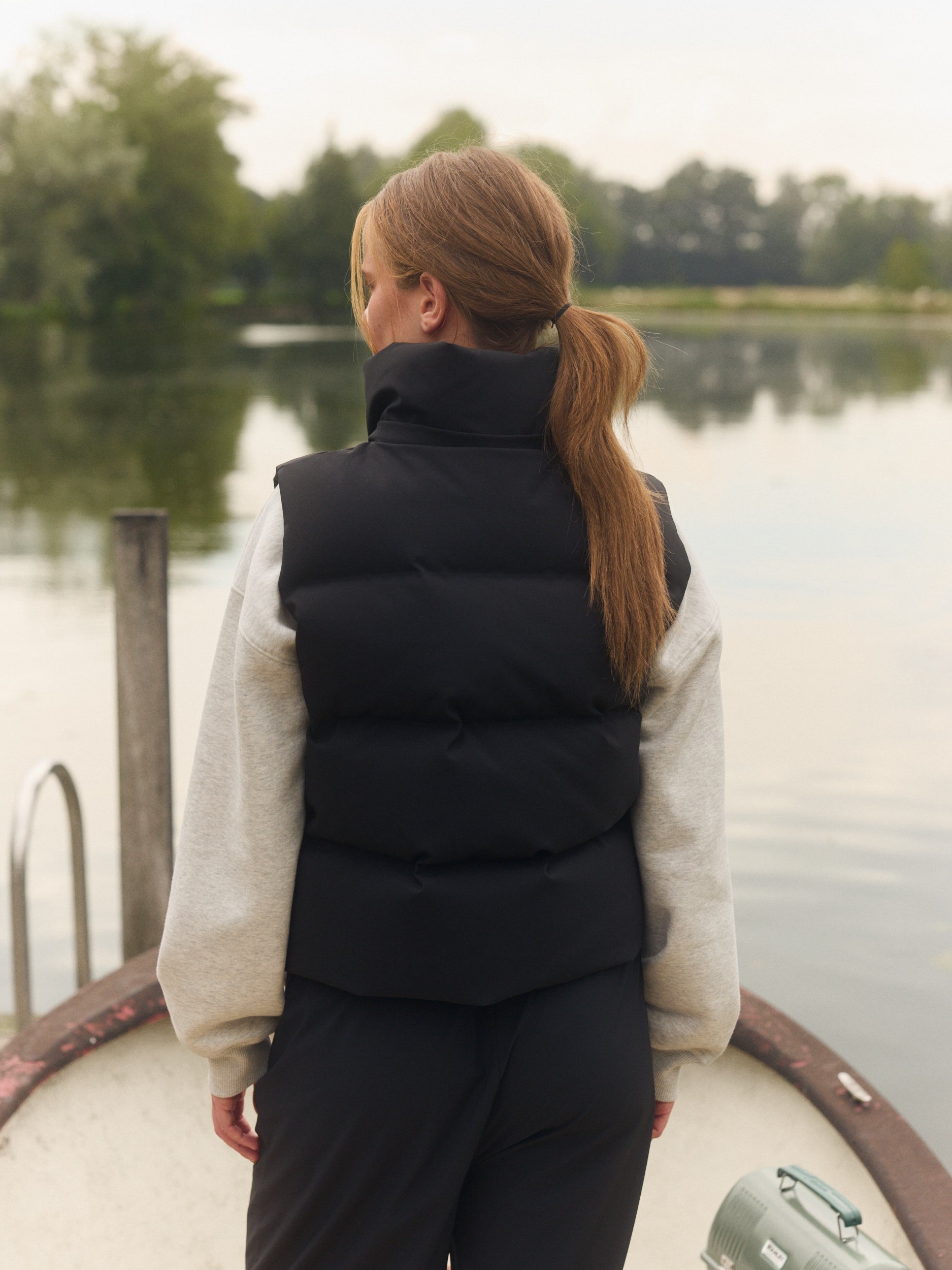 Back view of a woman wearing a black puffer vest over a grey sweatshirt by a lake.
