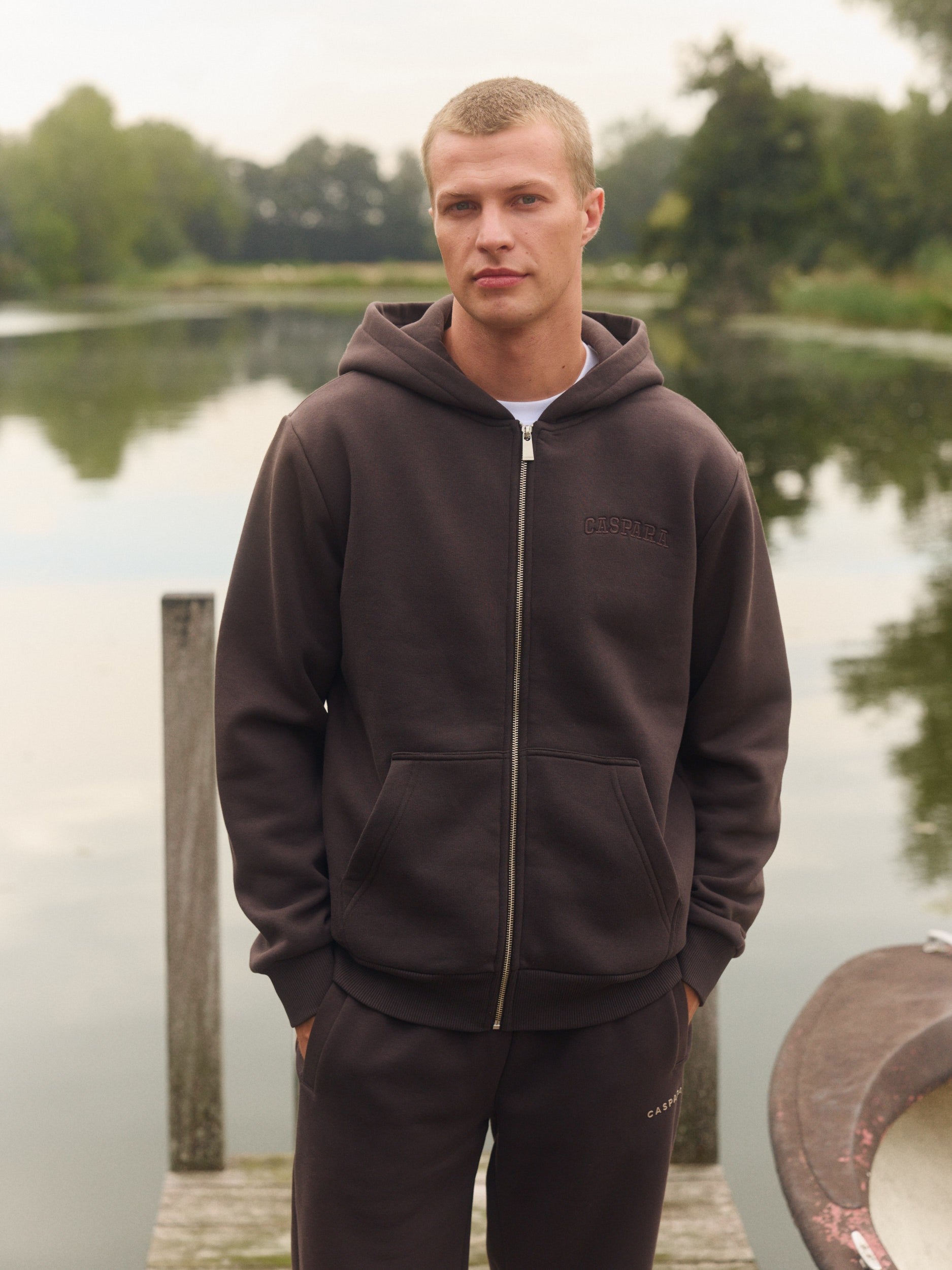 Man wearing an espresso brown zip-up hoodie standing on a wooden dock by a lake.
