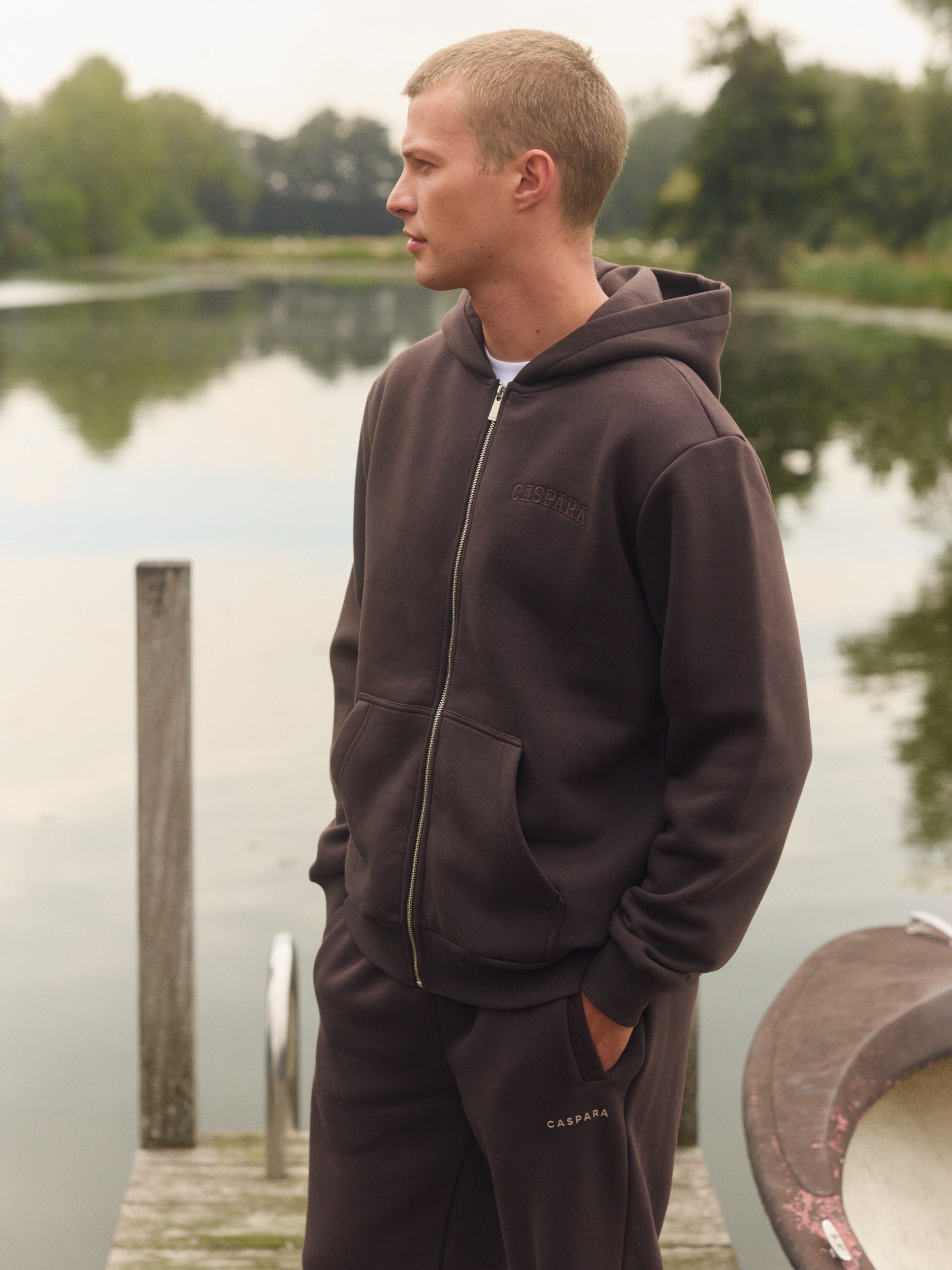 Man wearing an espresso brown zip-up hoodie standing on a wooden dock by a lake.