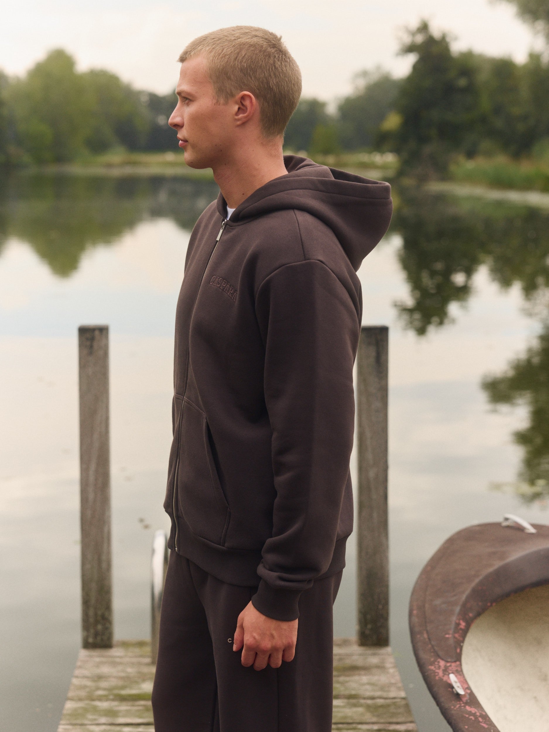 Man wearing an espresso brown zip hoodie standing on a wooden dock by a lake.
