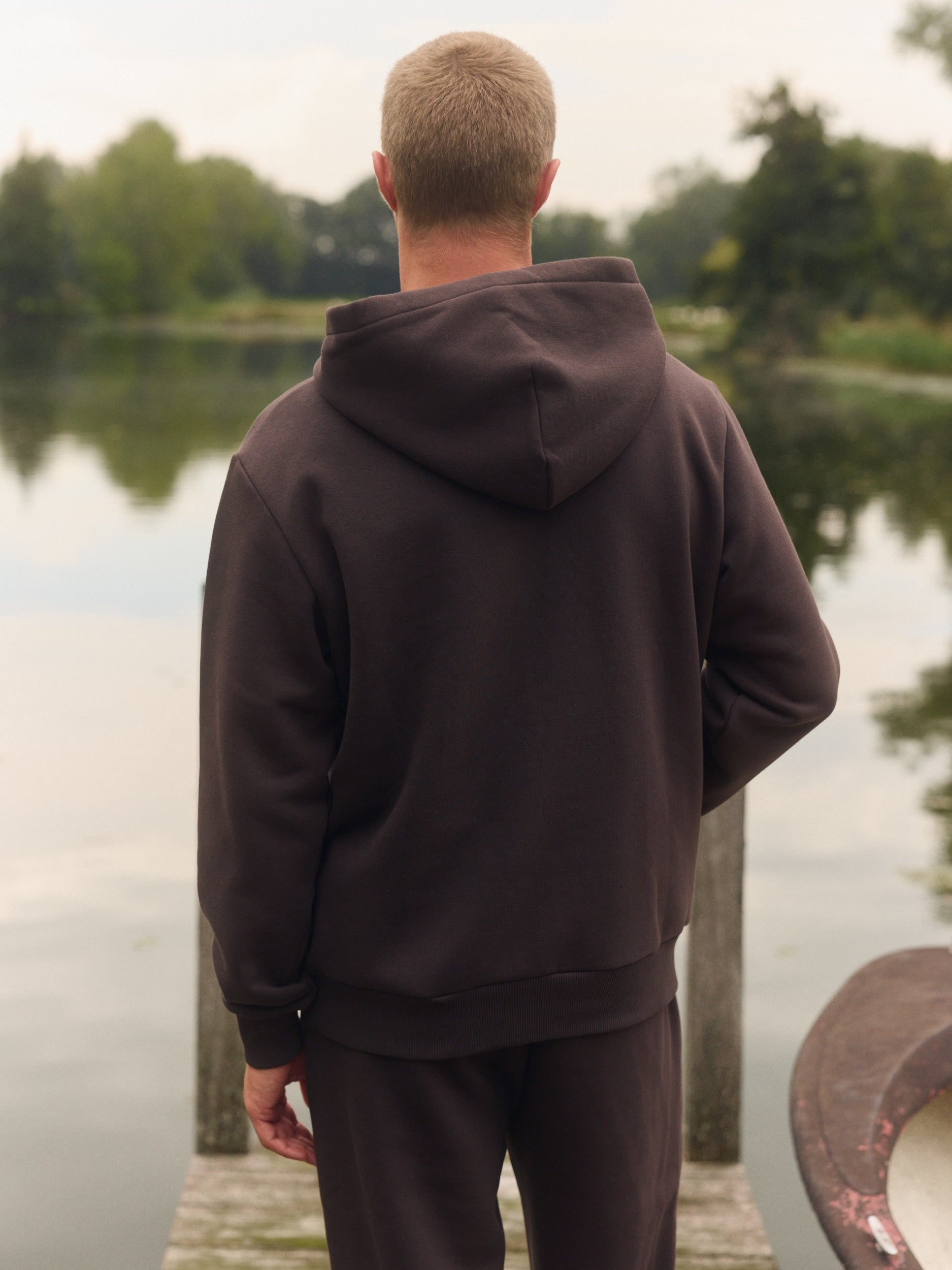 Man wearing an espresso brown hoodie seen from behind on a wooden dock by a lake.
