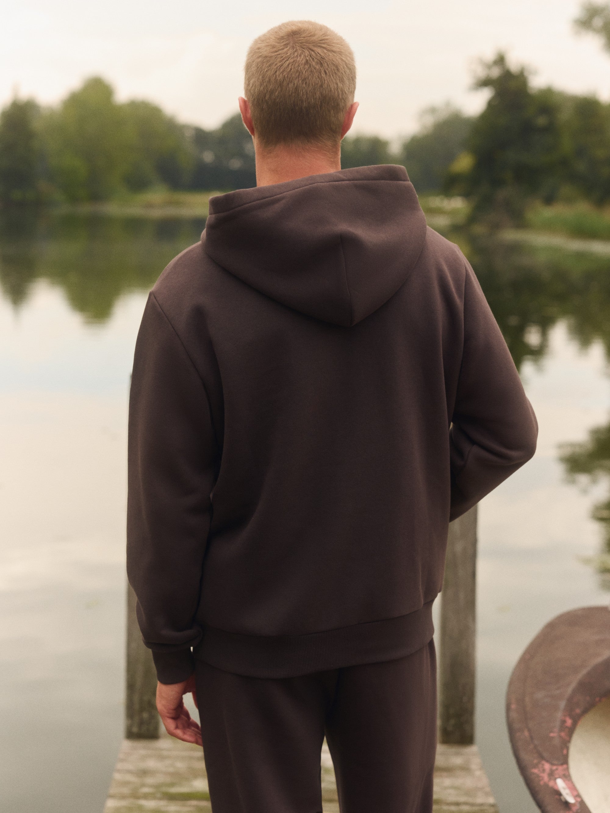 Back view of a man wearing an espresso brown CASPARA hoodie on a wooden dock by a lake.