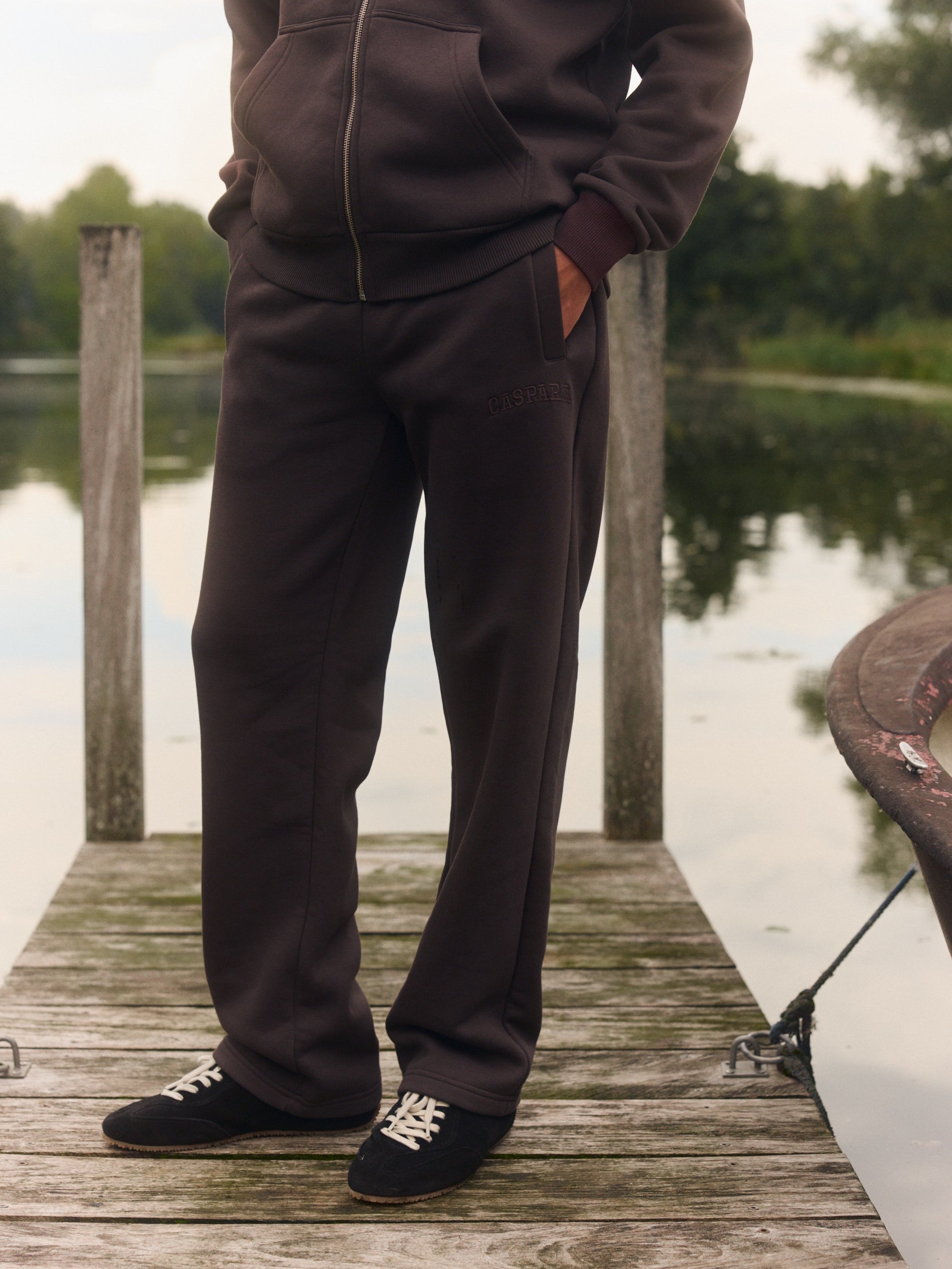 Man wearing espresso brown straight-leg sweatpants standing on a wooden dock by a lake.