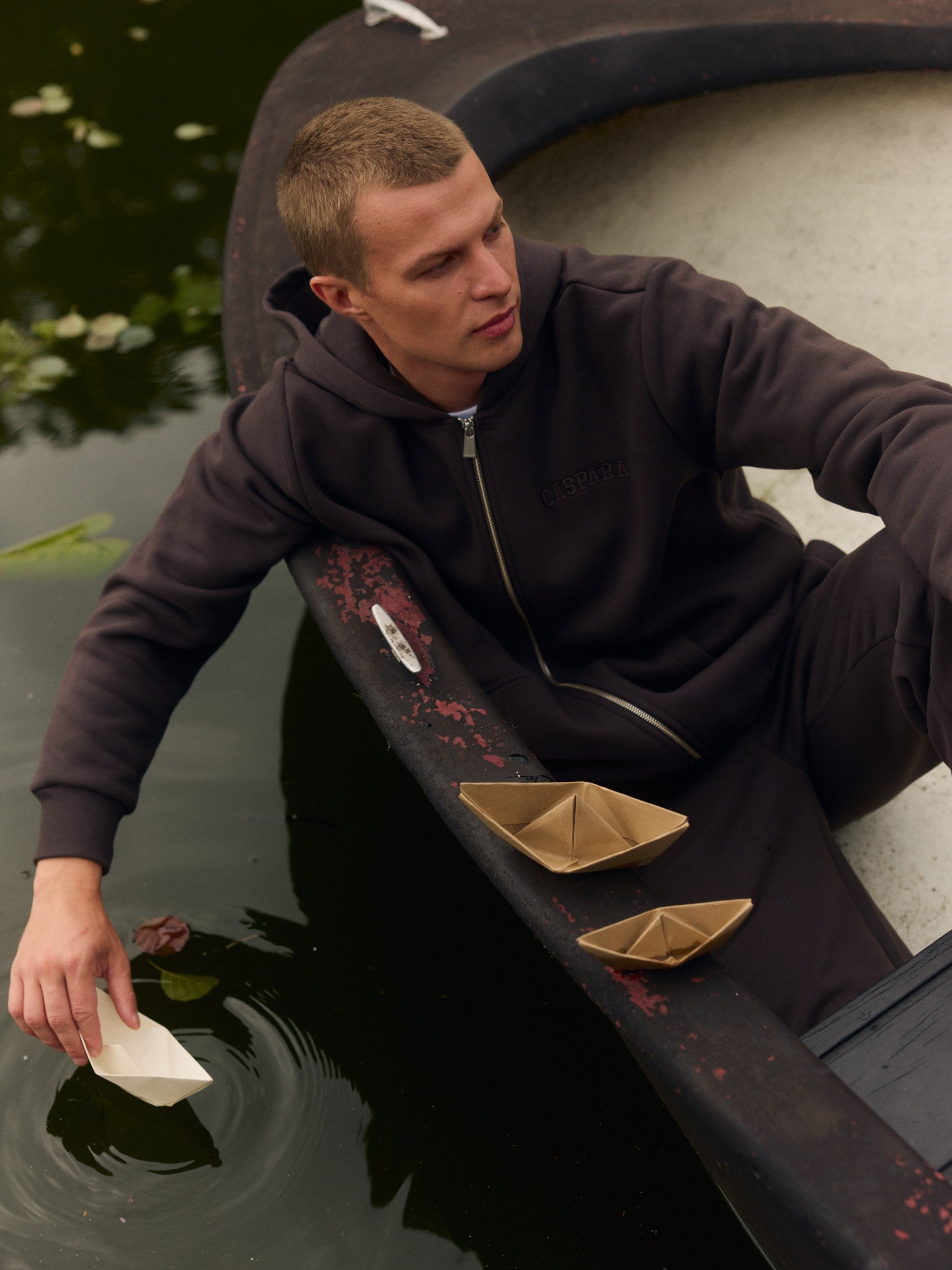 Man wearing an espresso zip hoodie sitting in a boat and placing a paper boat in the water.
