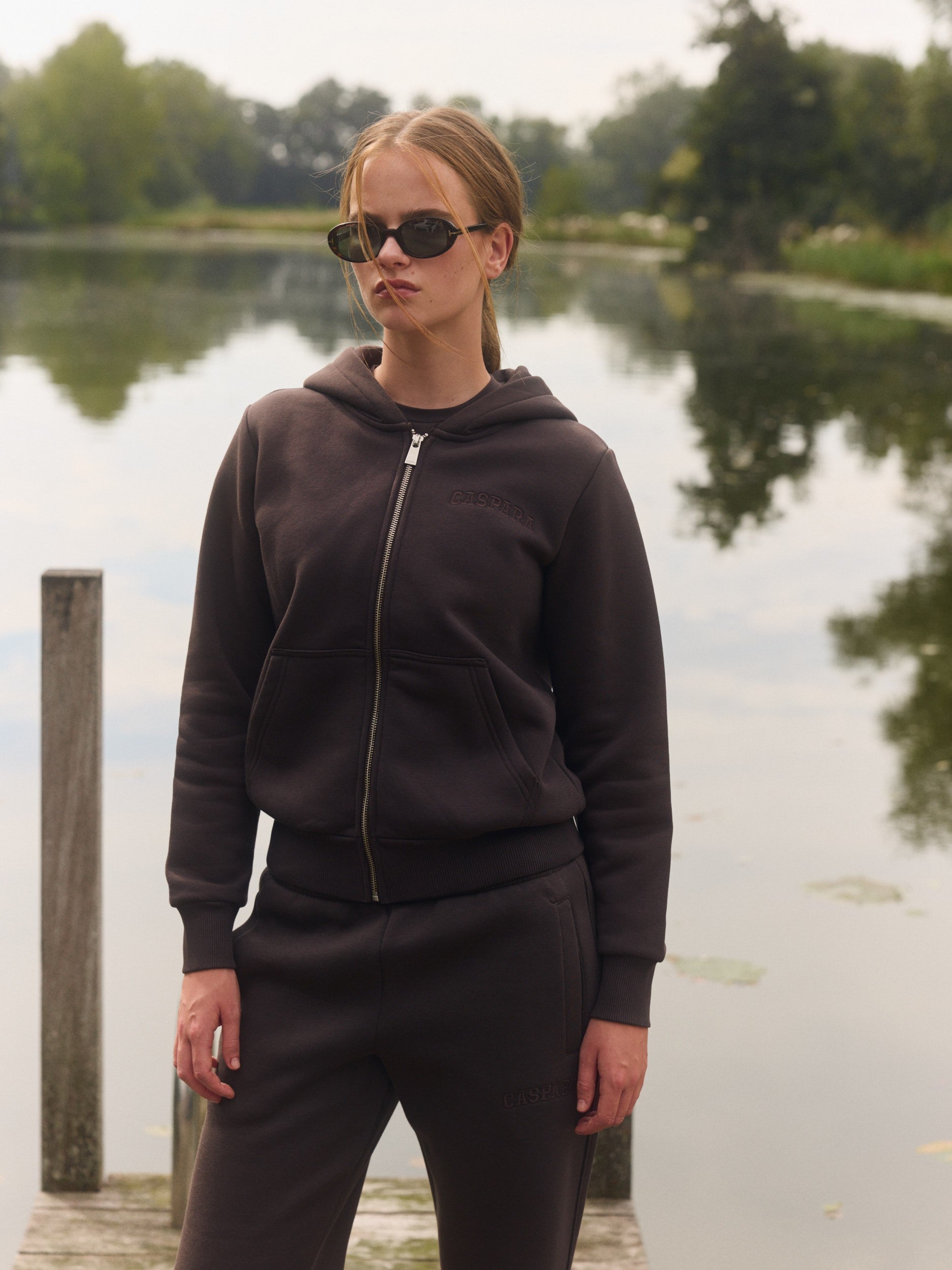 Woman wearing an espresso brown zip hoodie and sunglasses on a dock by a lake.