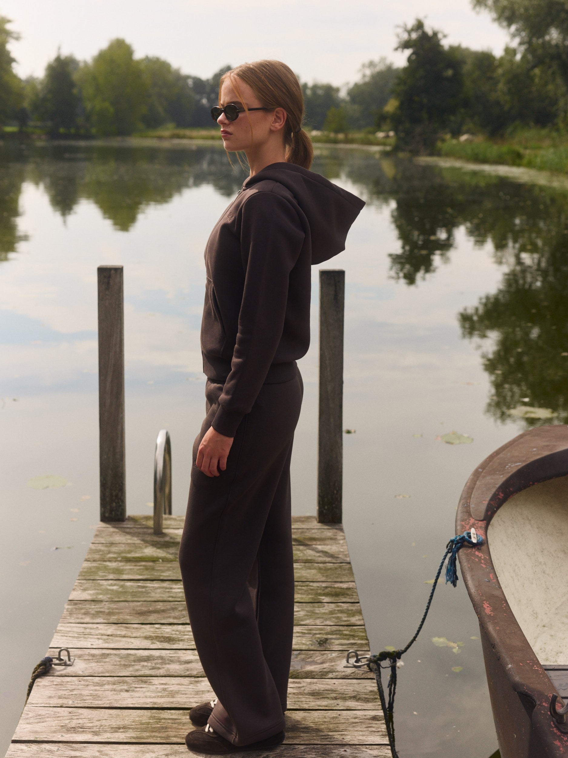 Woman wearing an espresso zip hoodie and matching sweatpants on a wooden dock by a lake.