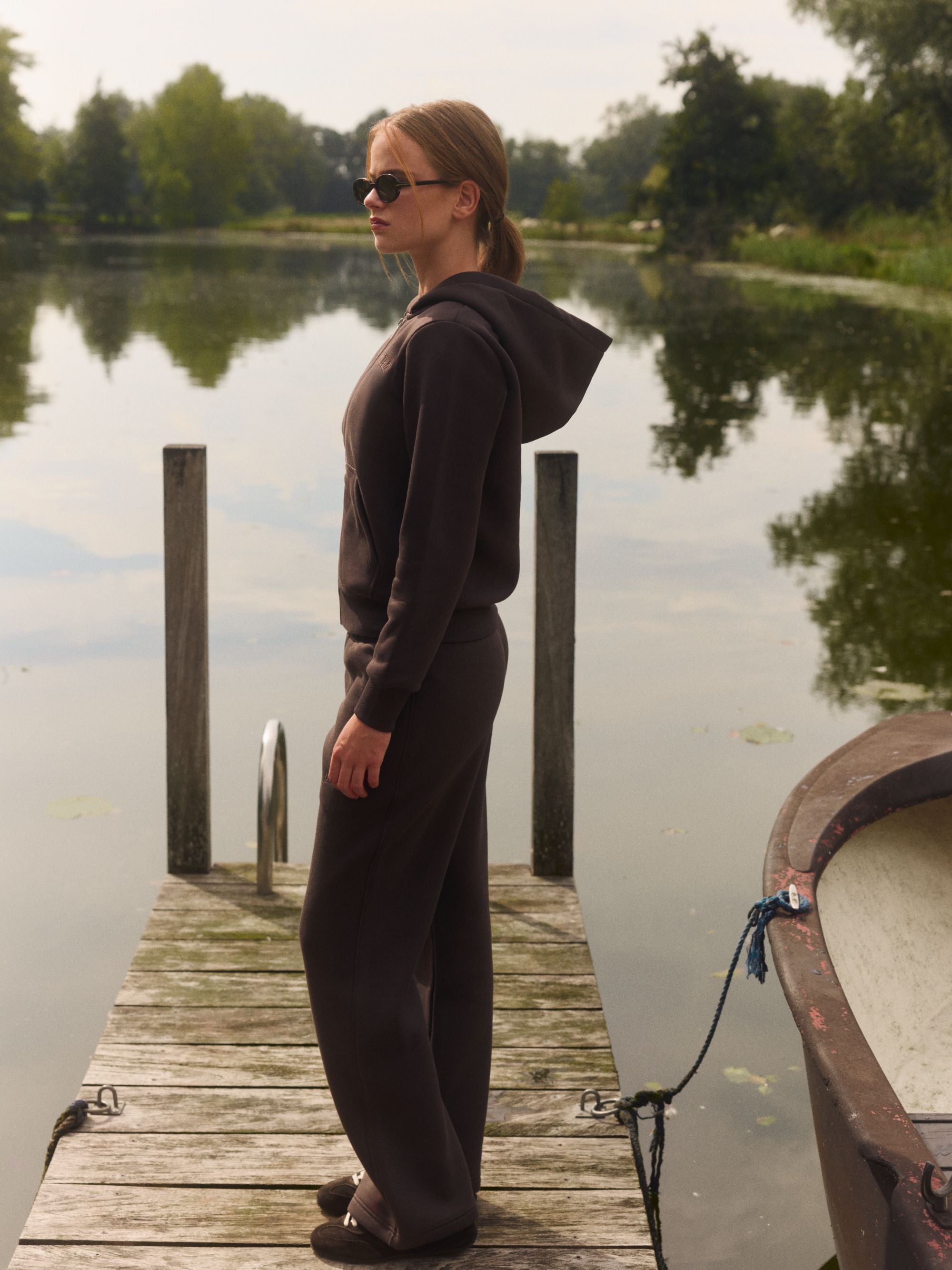 Woman wearing an espresso CASPARA zip hoodie and sunglasses on a wooden dock by a lake.