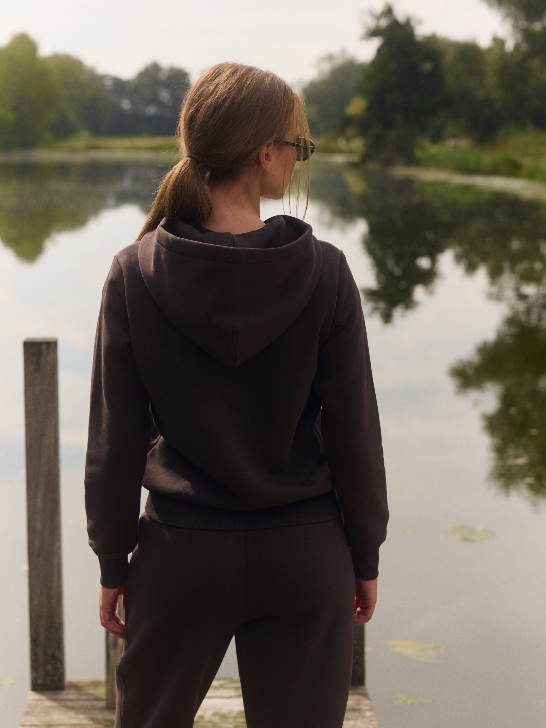 Woman wearing an espresso brown zip hoodie standing on a wooden dock by a calm lake.