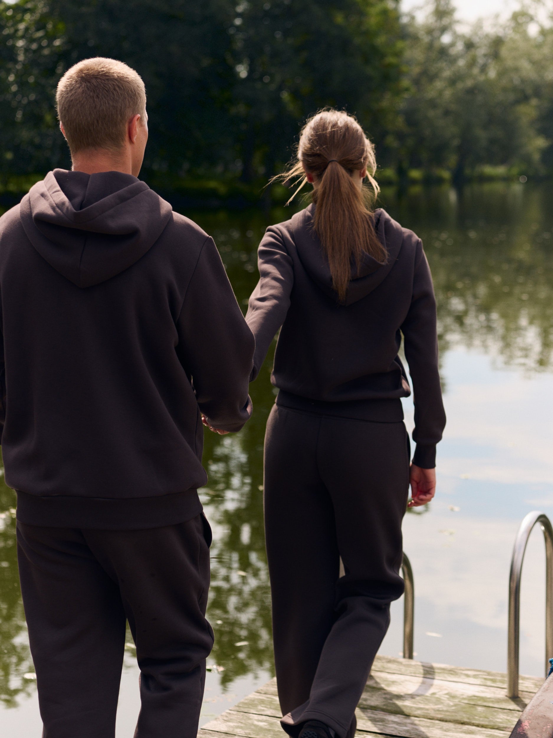 Couple wearing matching espresso brown hoodies and sweatpants on a wooden dock by a lake.