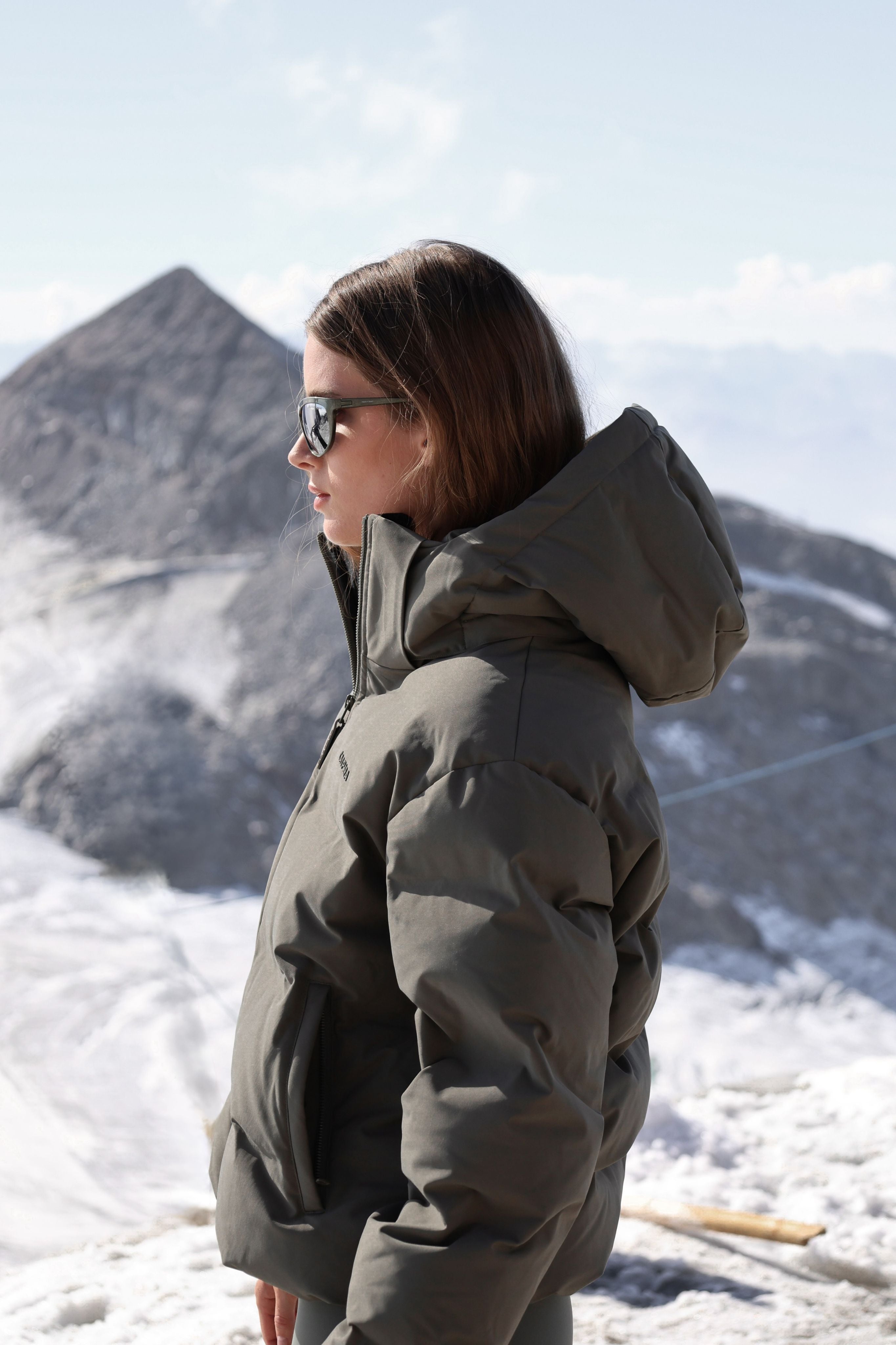 Woman wearing a khaki CASPARA Aurora puffer jacket in a snowy mountain setting.