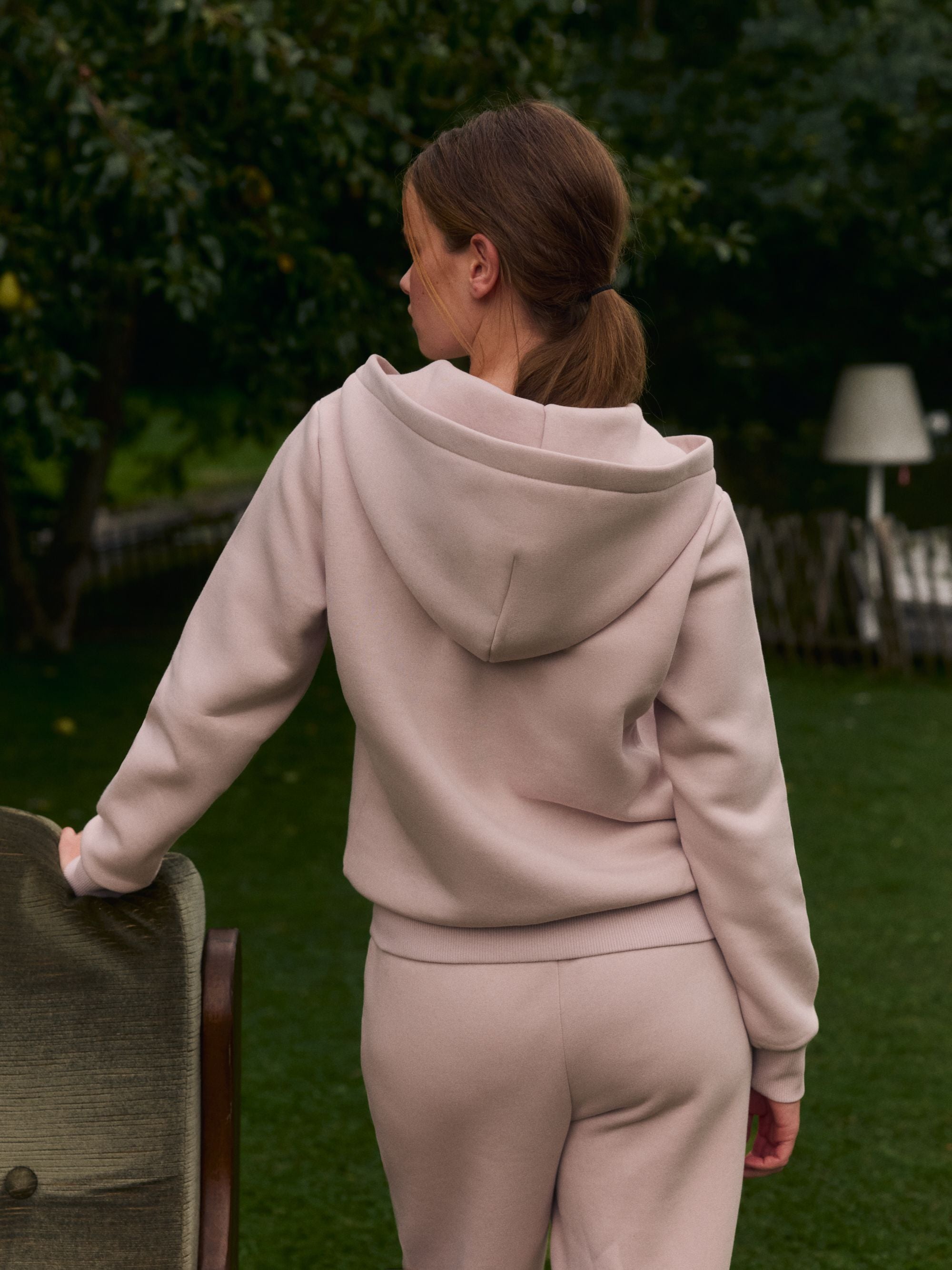 Woman wearing a dusty pink CASPARA zip hoodie and matching sweatpants outdoors in a garden.