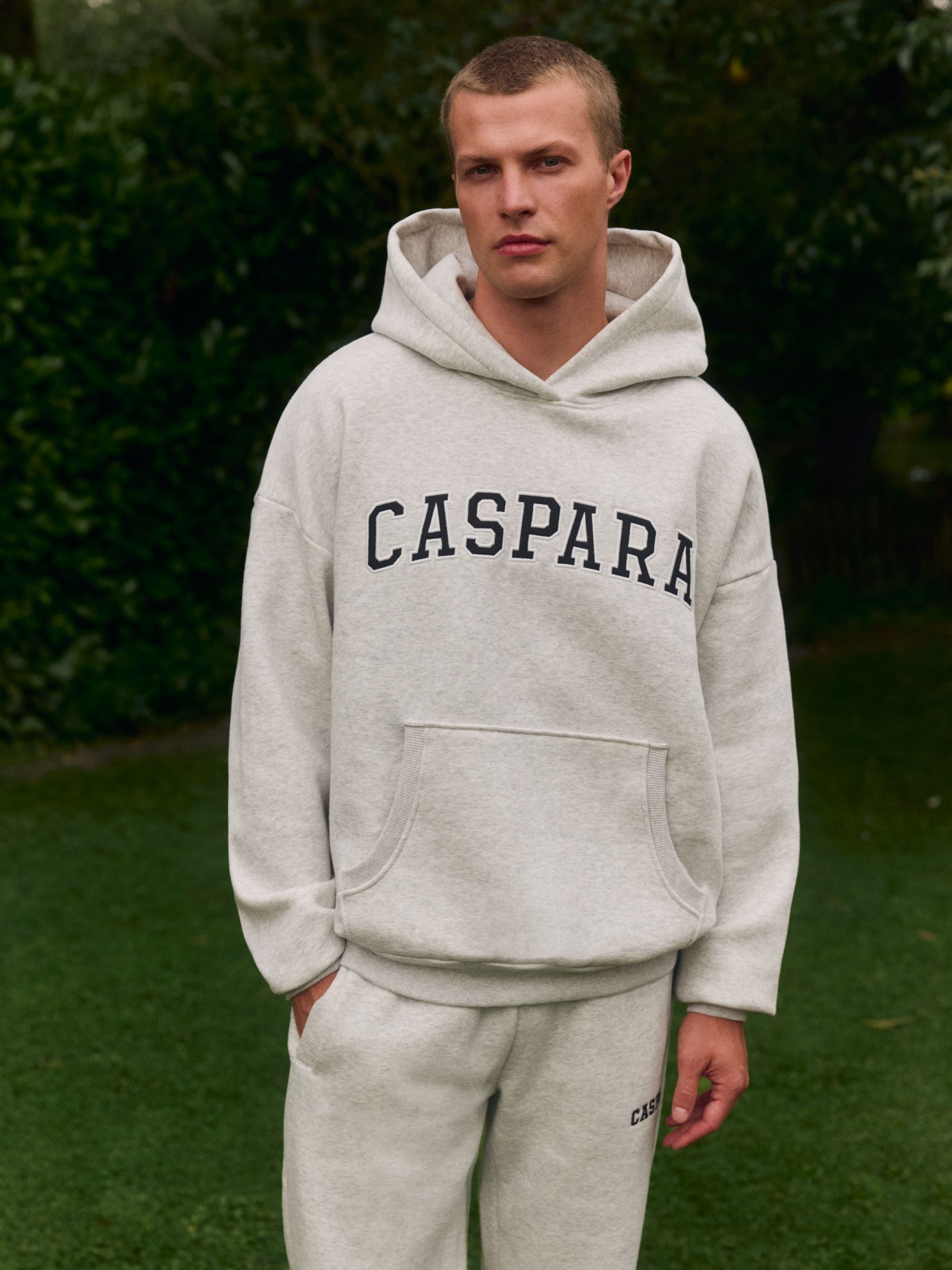 Man wearing a grey melange CASPARA logo hoodie and matching sweatpants outdoors.