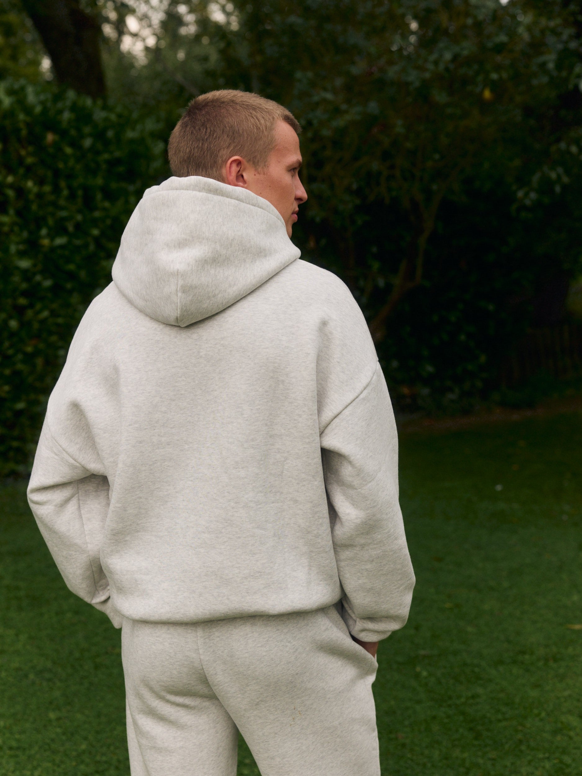 Back view of a man wearing a grey melange hoodie and matching sweatpants in a garden.