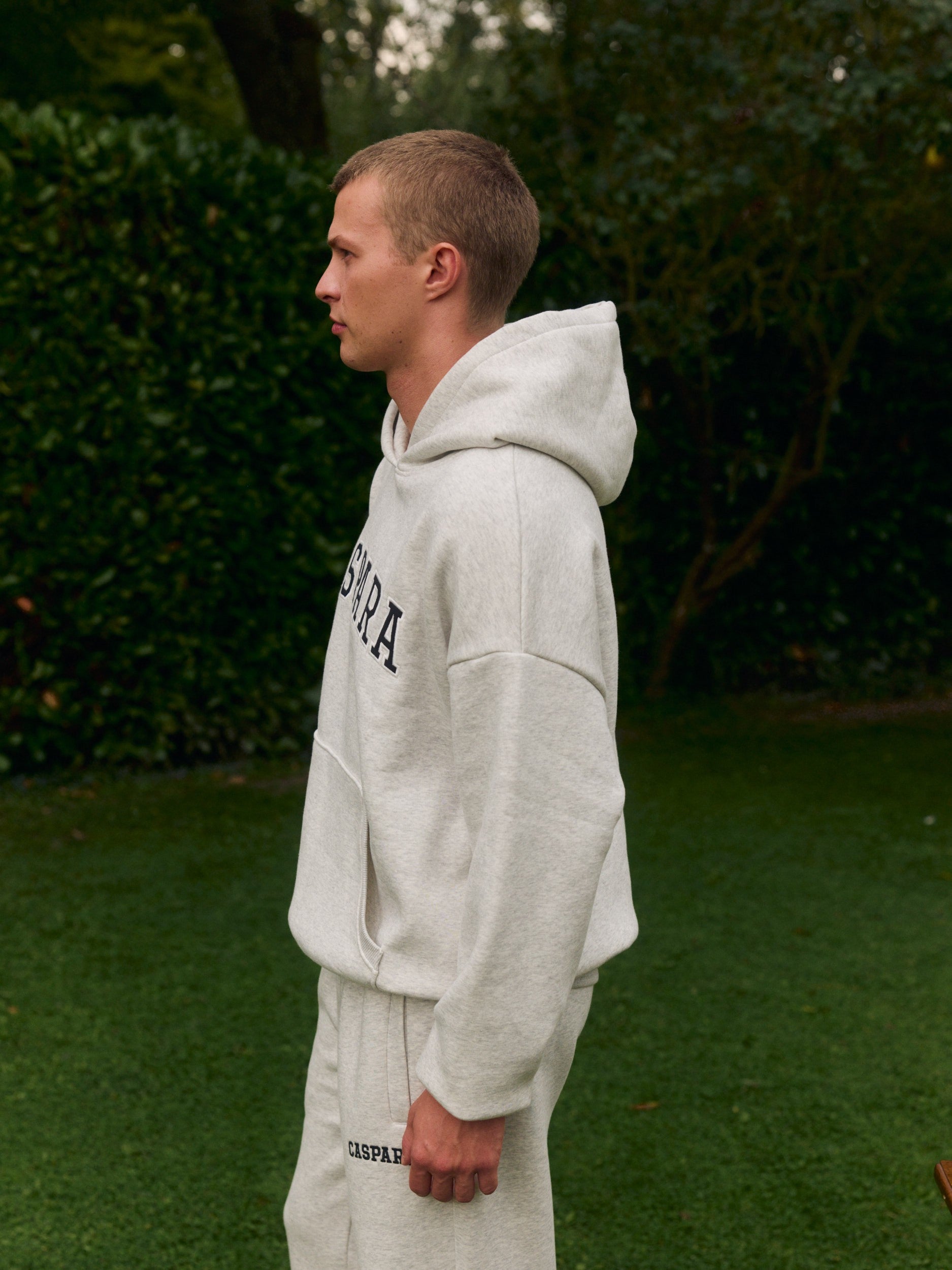 Man wearing a grey melange hoodie standing outdoors in front of green foliage.