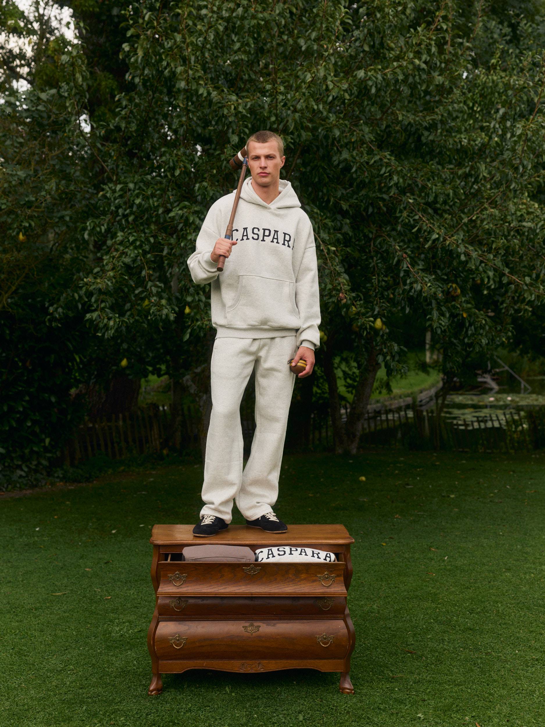 Man in a grey melange hoodie and sweatpants standing on a wooden dresser outdoors with a croquet mallet.