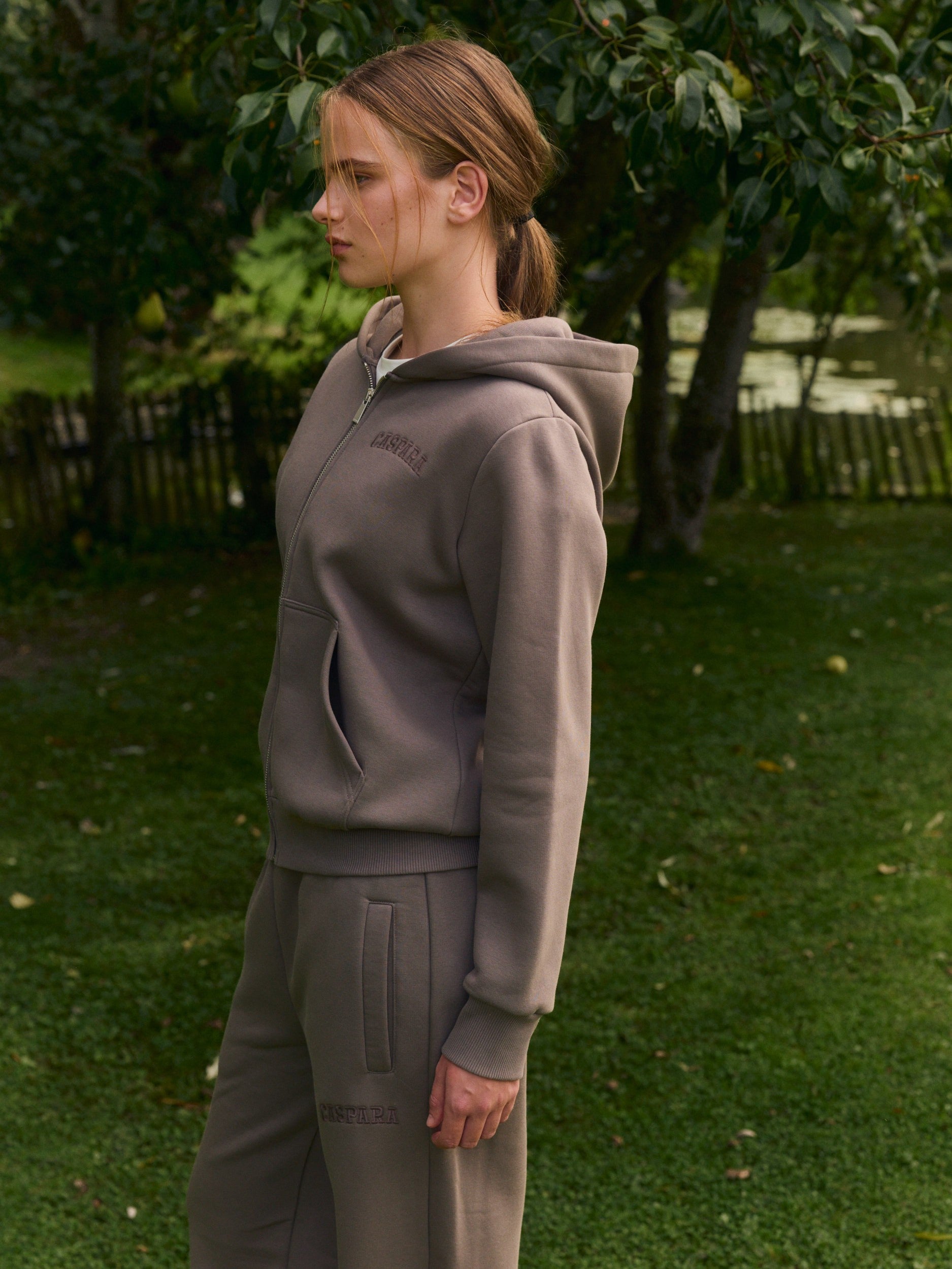 Woman wearing a walnut zip hoodie and matching sweatpants standing outdoors in a garden.
