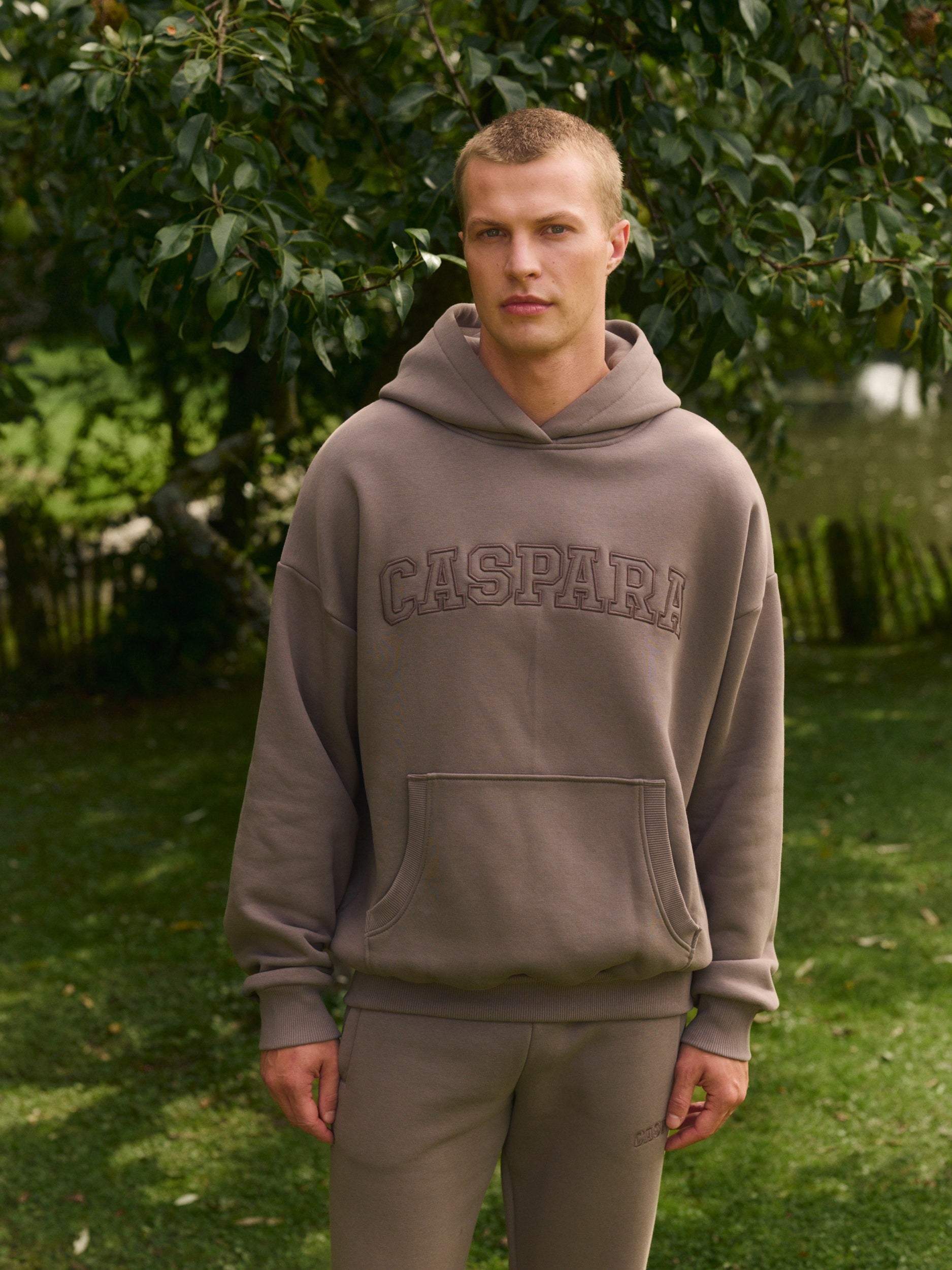 Man wearing a walnut brown CASPARA hoodie and matching sweatpants in a garden.