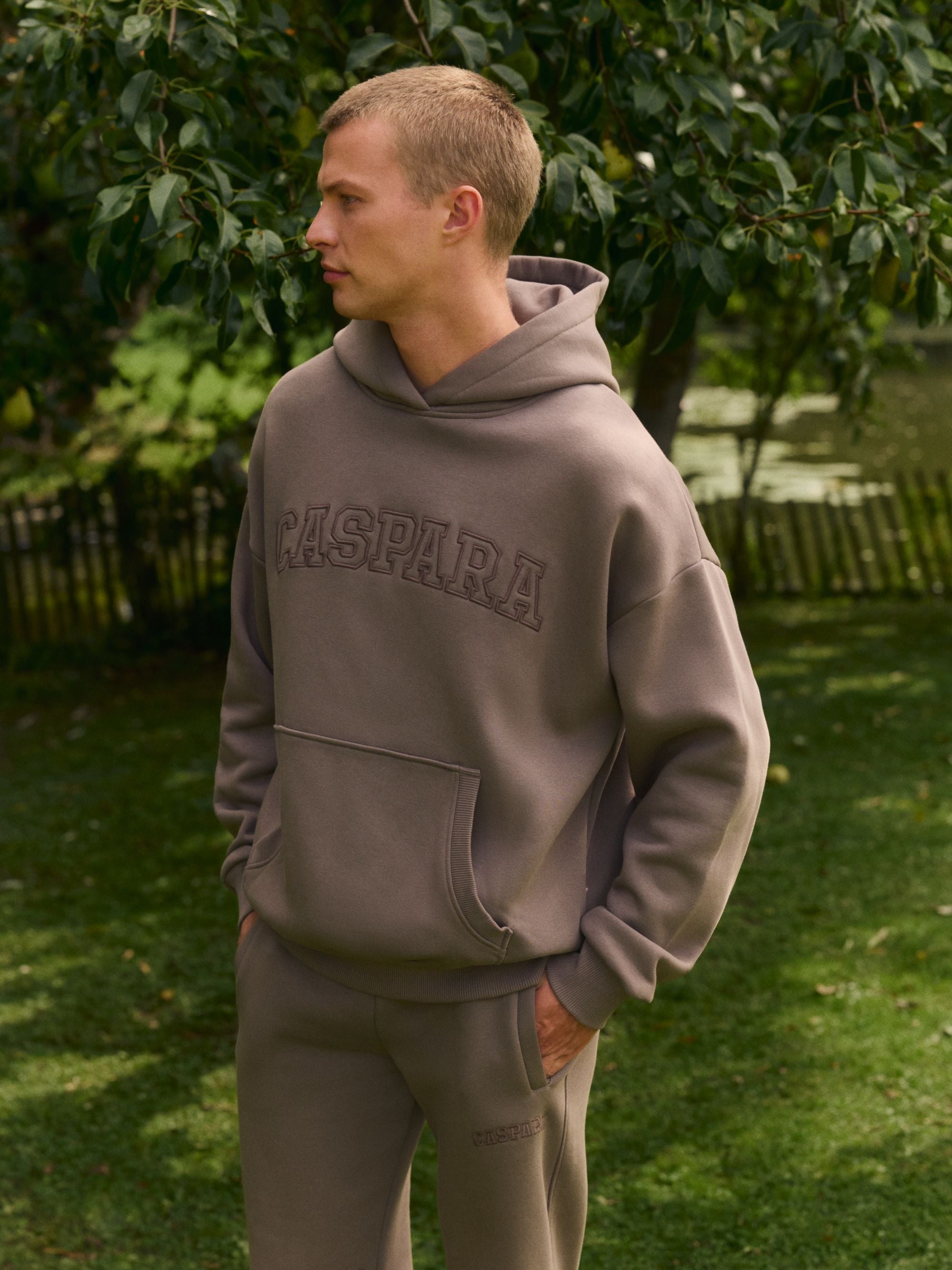Man wearing a walnut CASPARA logo hoodie and matching sweatpants in an outdoor garden setting.