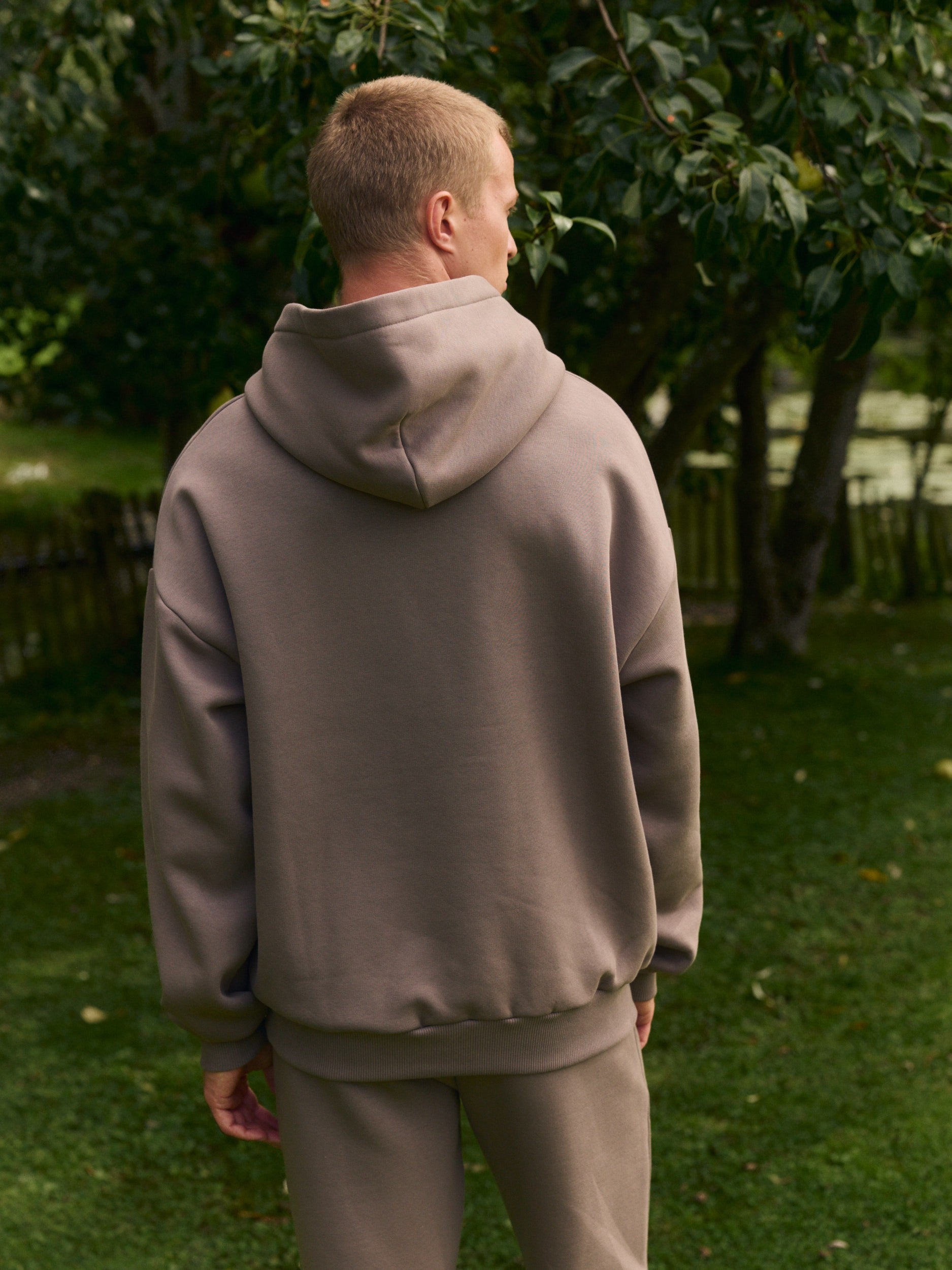Back view of a man wearing a walnut oversized hoodie in an outdoor garden.