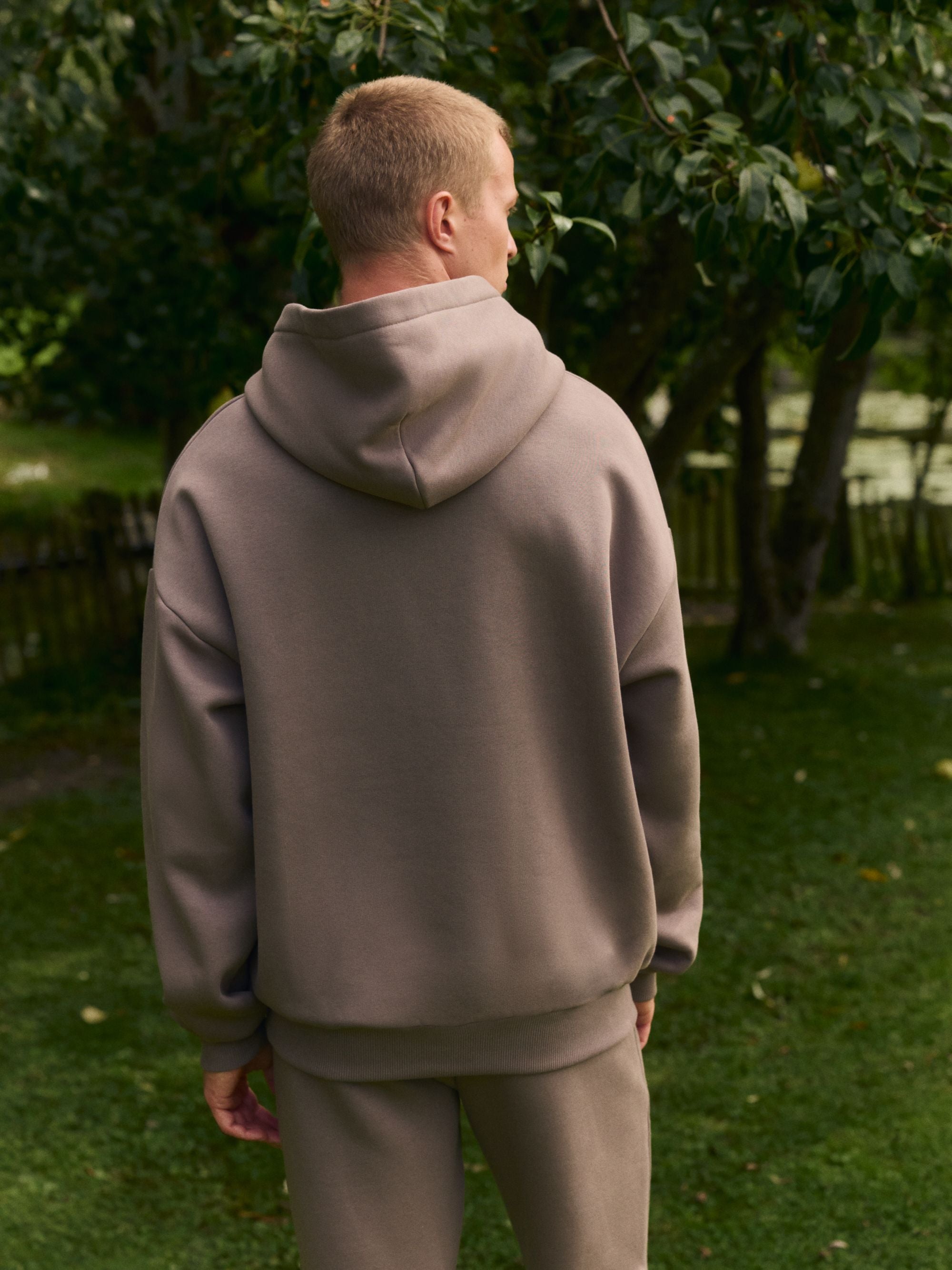 Back view of a man wearing a walnut CASPARA hoodie and matching pants outdoors.