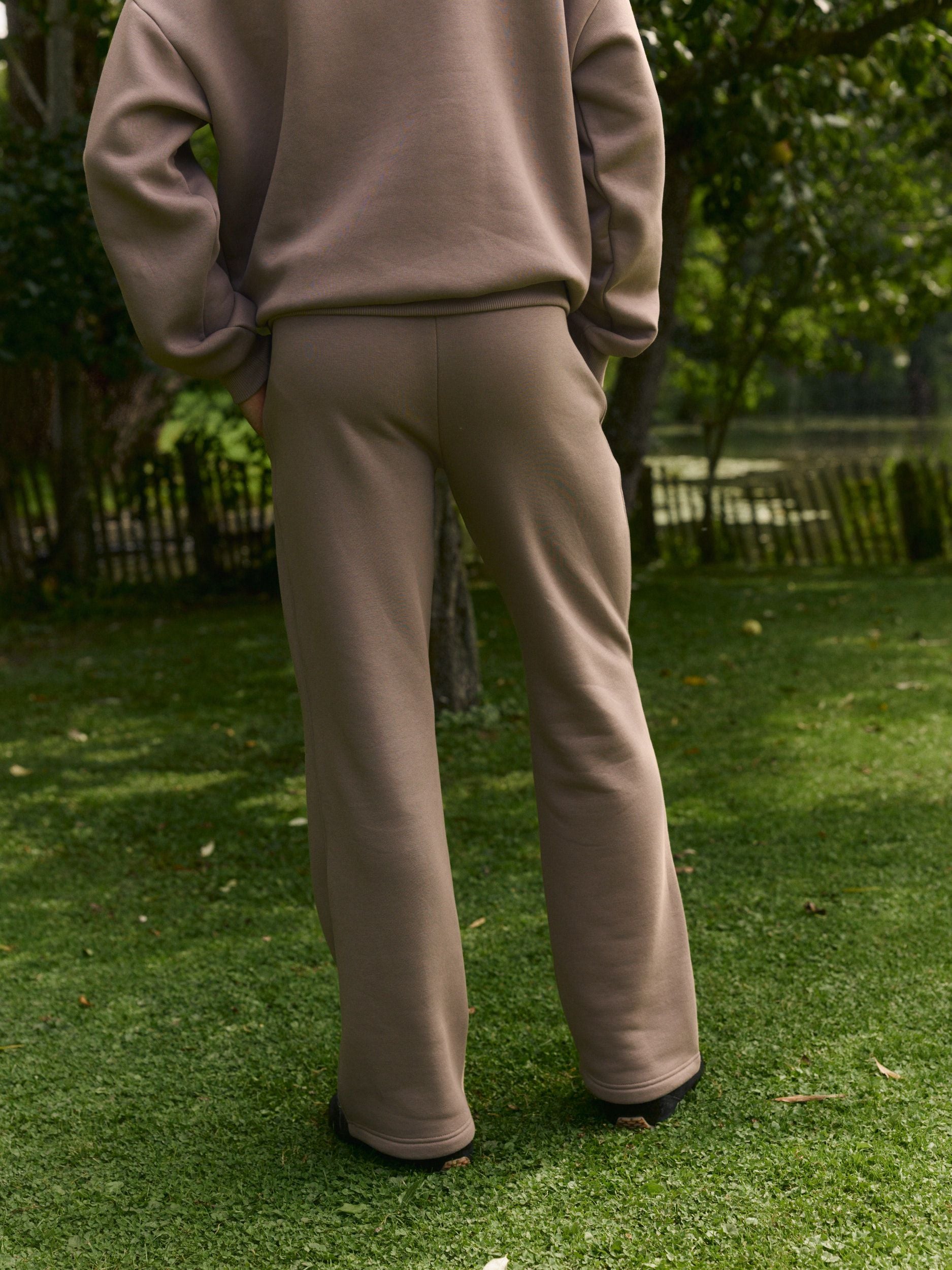 Back view of a man wearing walnut straight-leg sweatpants standing outdoors on grass.