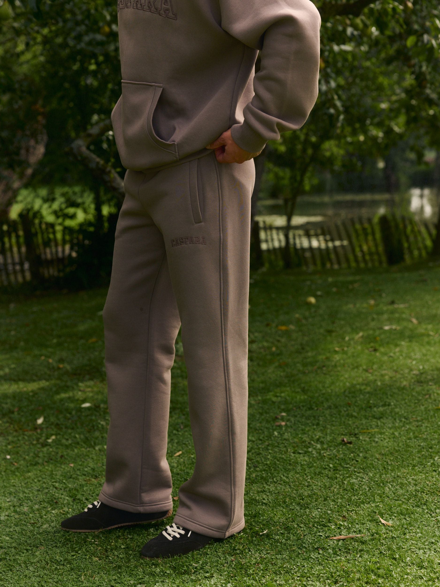 Man wearing walnut straight-leg sweatpants and matching hoodie standing on a green lawn.