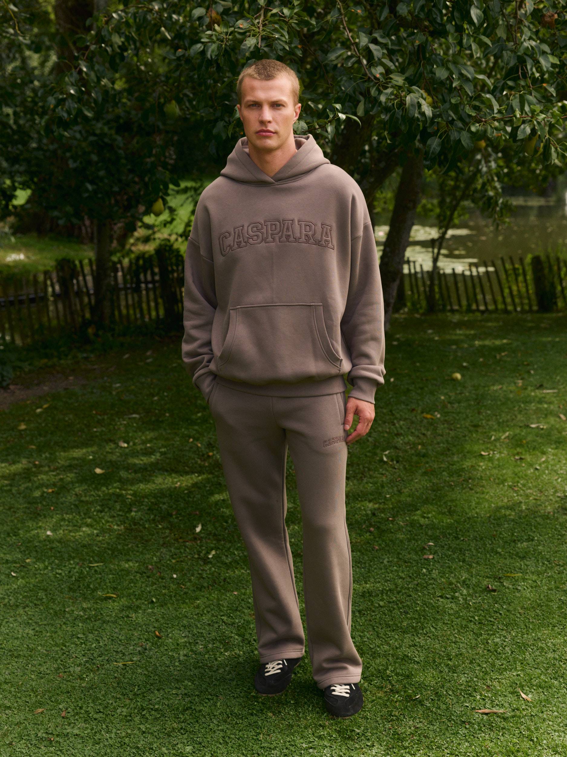Man wearing a walnut brown hoodie and matching straight-leg sweatpants on a grassy lawn.