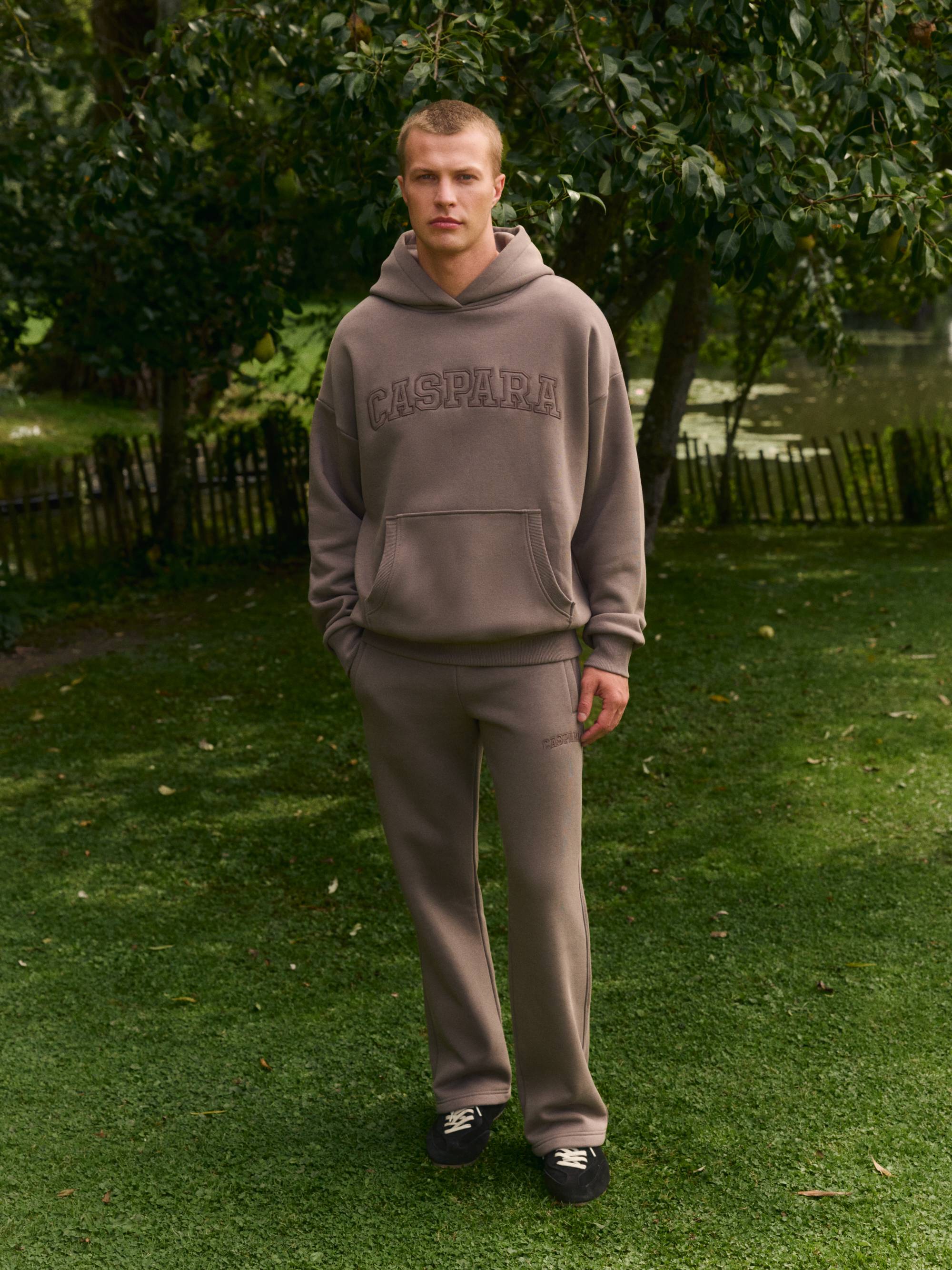 Man wearing a walnut CASPARA logo hoodie and matching straight-leg sweatpants outdoors.