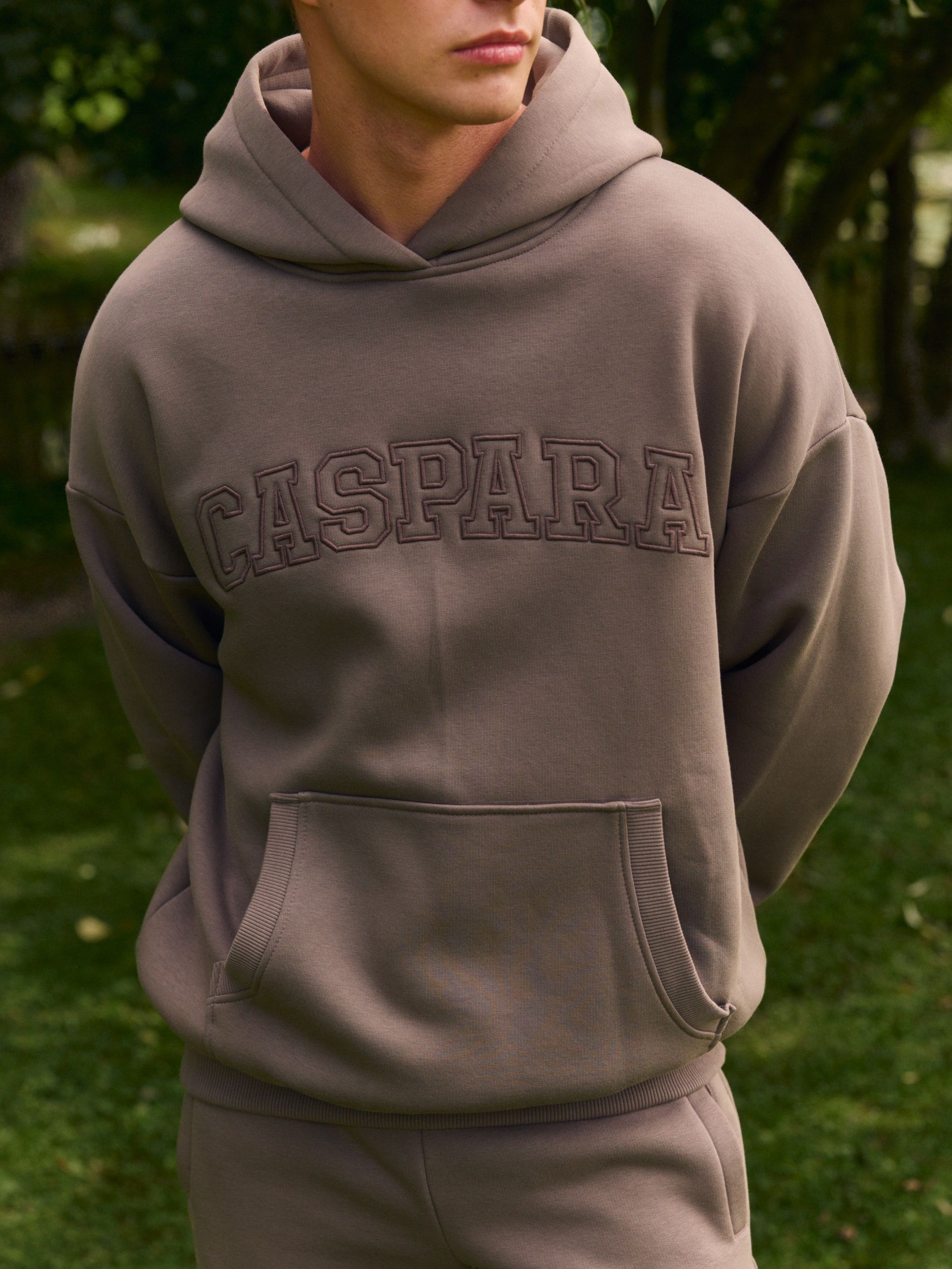 Man wearing a walnut brown hoodie with embroidered CASPARA logo outdoors.