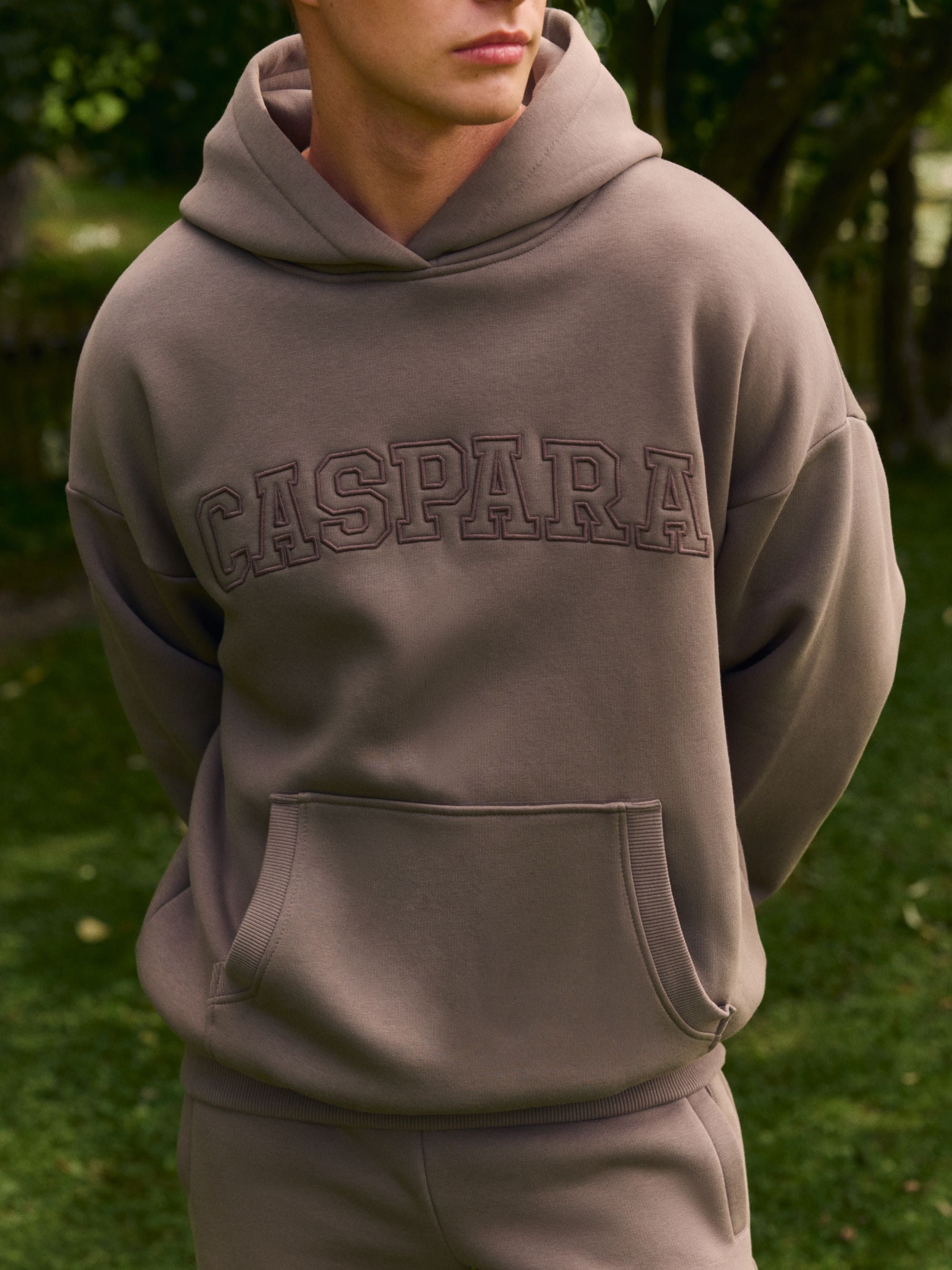 Man wearing a walnut CASPARA embroidered logo hoodie outdoors.