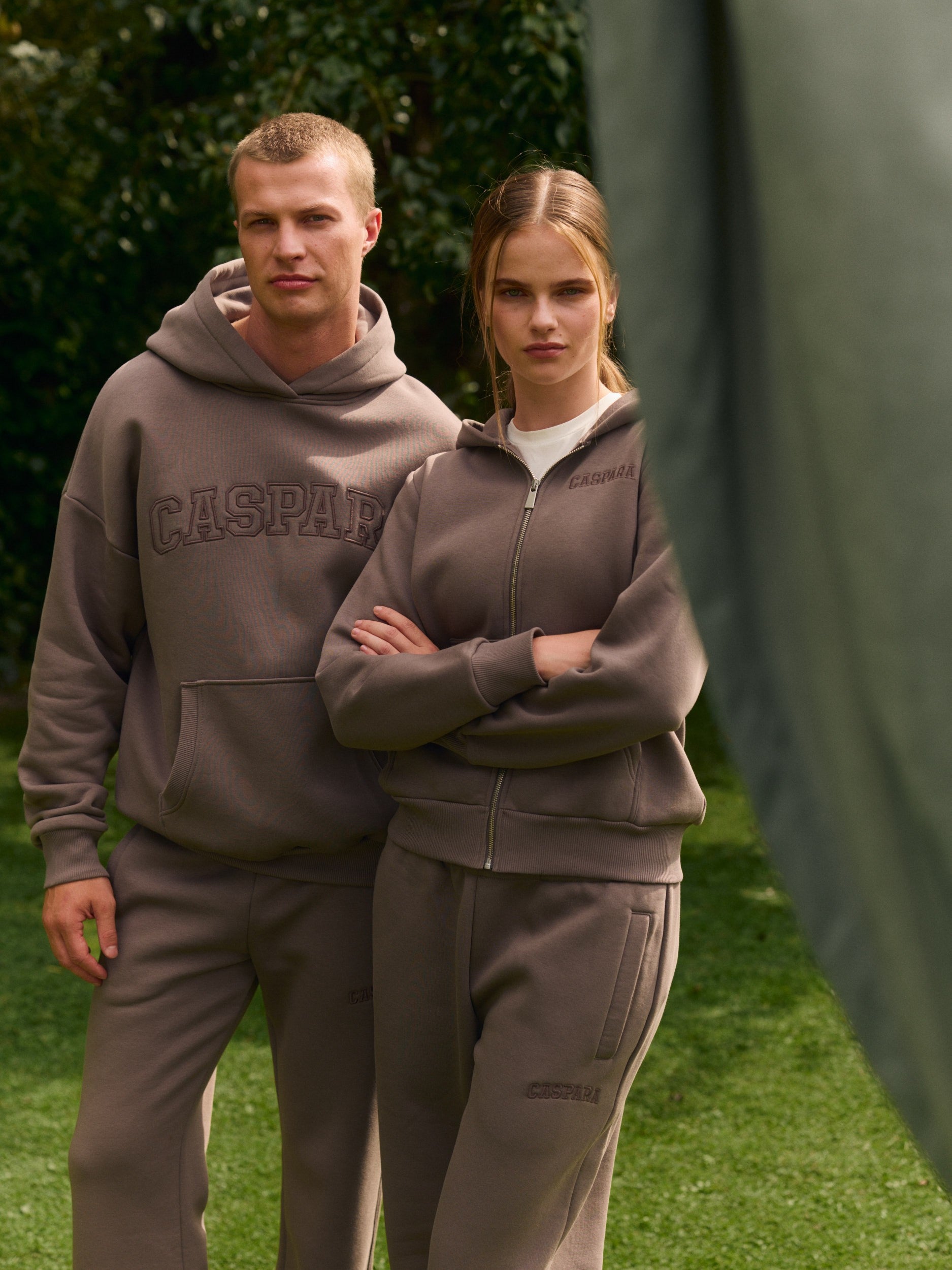 Man wearing a walnut CASPARA hoodie and sweatpants beside a woman in a matching zip hoodie.