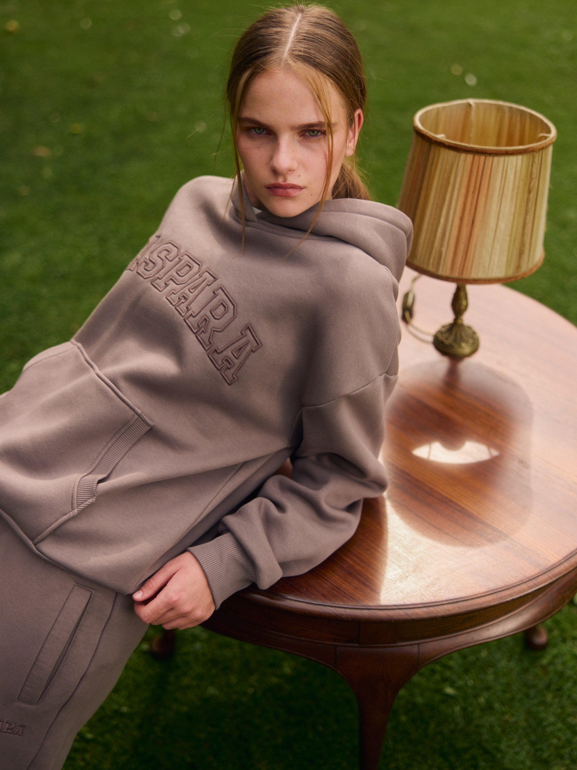 Woman wearing a walnut CASPARA hoodie leaning against a wooden table on a green lawn.