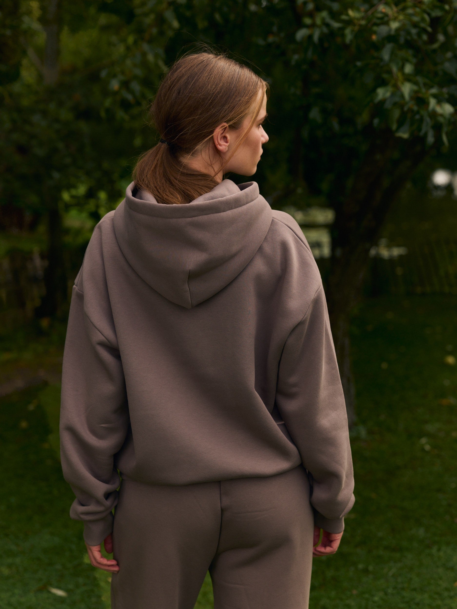 Woman viewed from behind wearing an oversized walnut hoodie in a garden setting.